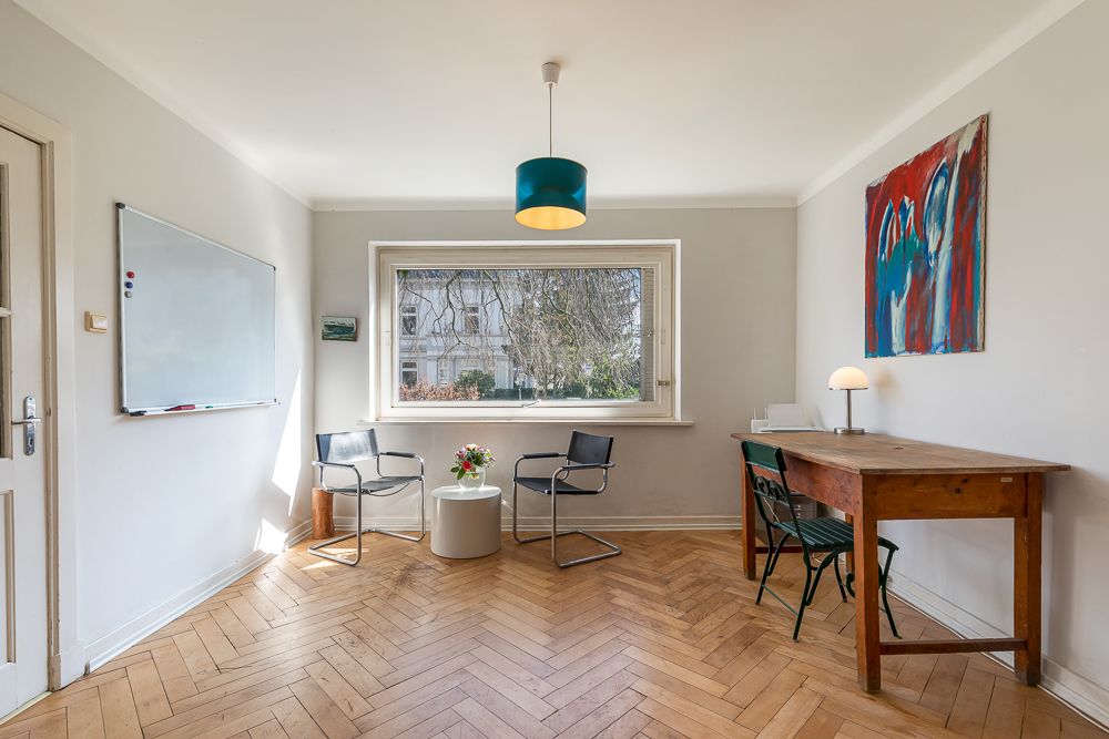 Heller Raum mit großem Fenster, zwei Stühlen, rundem Tisch, Schreibtisch und Wandtafel.