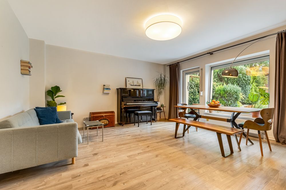 Helles Wohnzimmer mit Sofa, Klavier, Esstisch, Holzboden und großen Fenstern zum Garten.