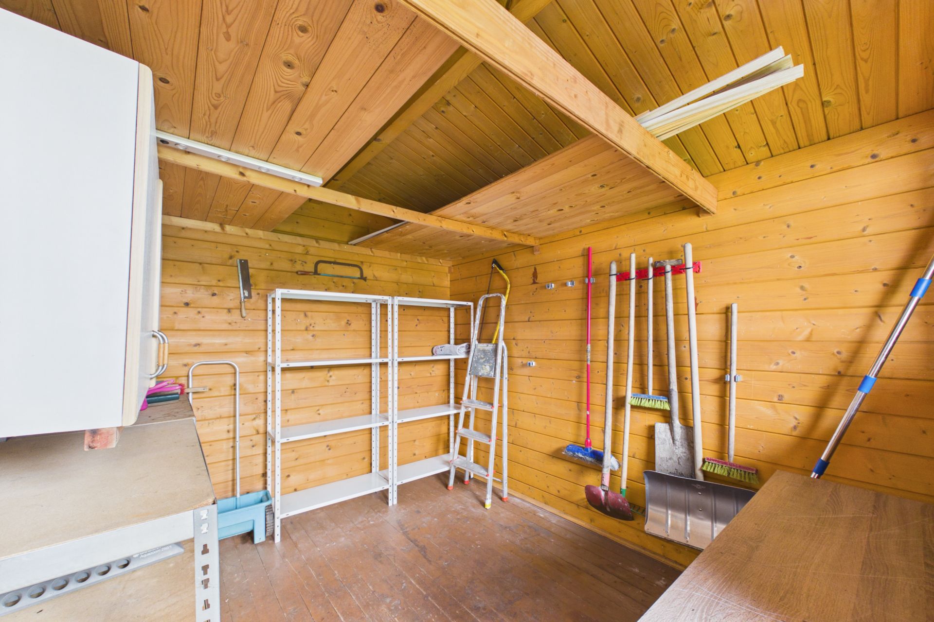Holzraum mit Regalen, Leitern und an der Wand hängenden Gartengeräten.