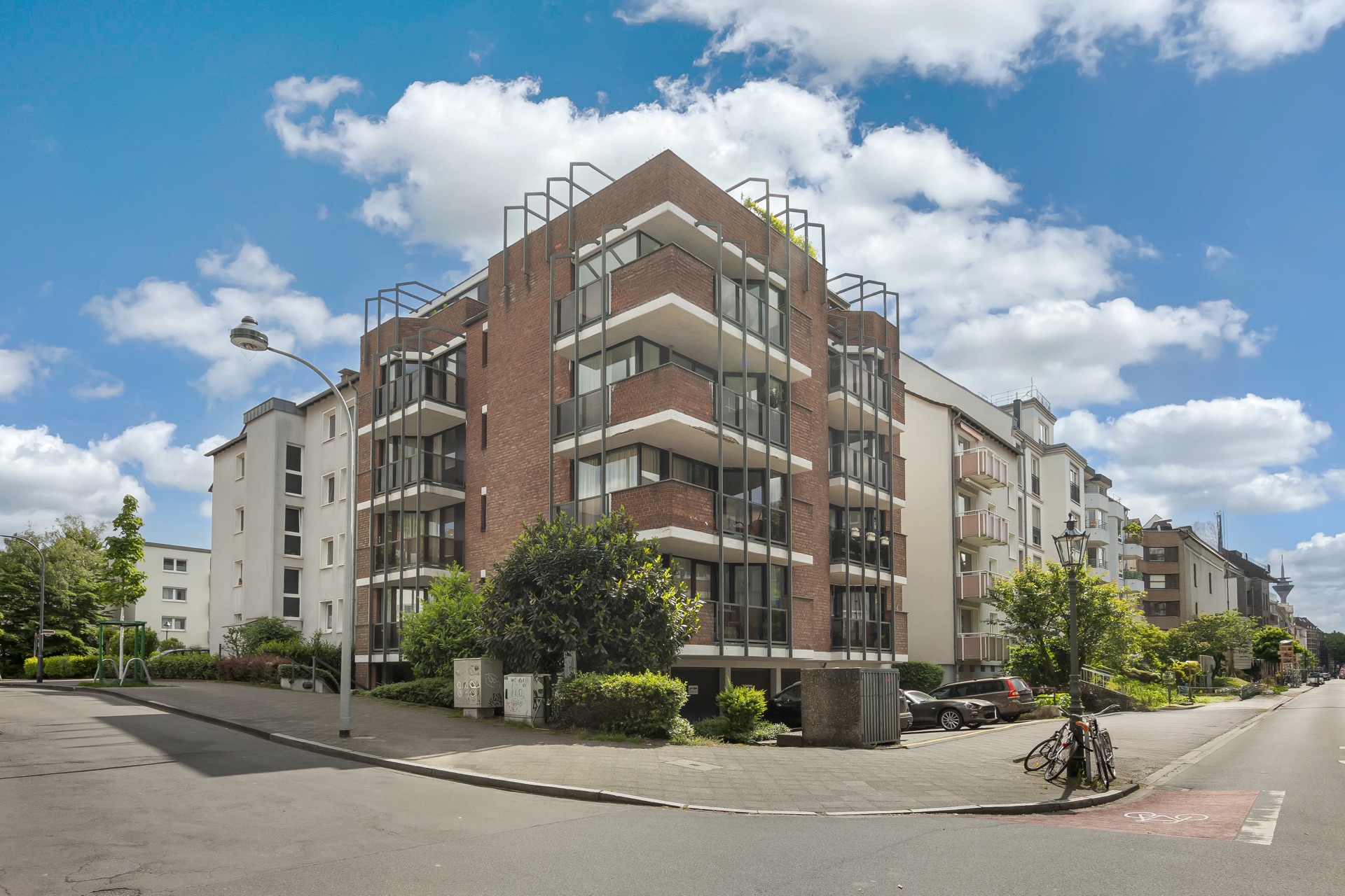 Mehrstöckiges Wohnhaus mit Balkonen an einer Straßenecke, umgeben von Bäumen und Gehweg.