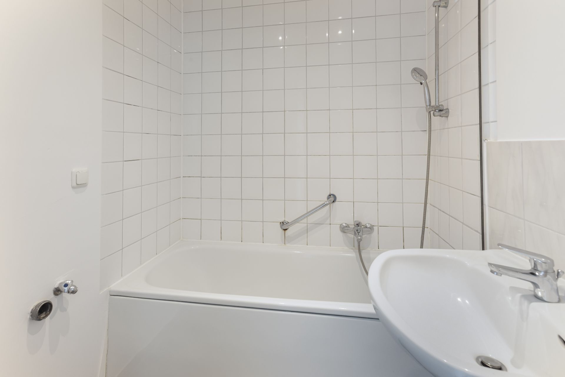 Weiße Badewanne mit Duschkopf und Waschbecken vor einer gefliesten Wand im Badezimmer.