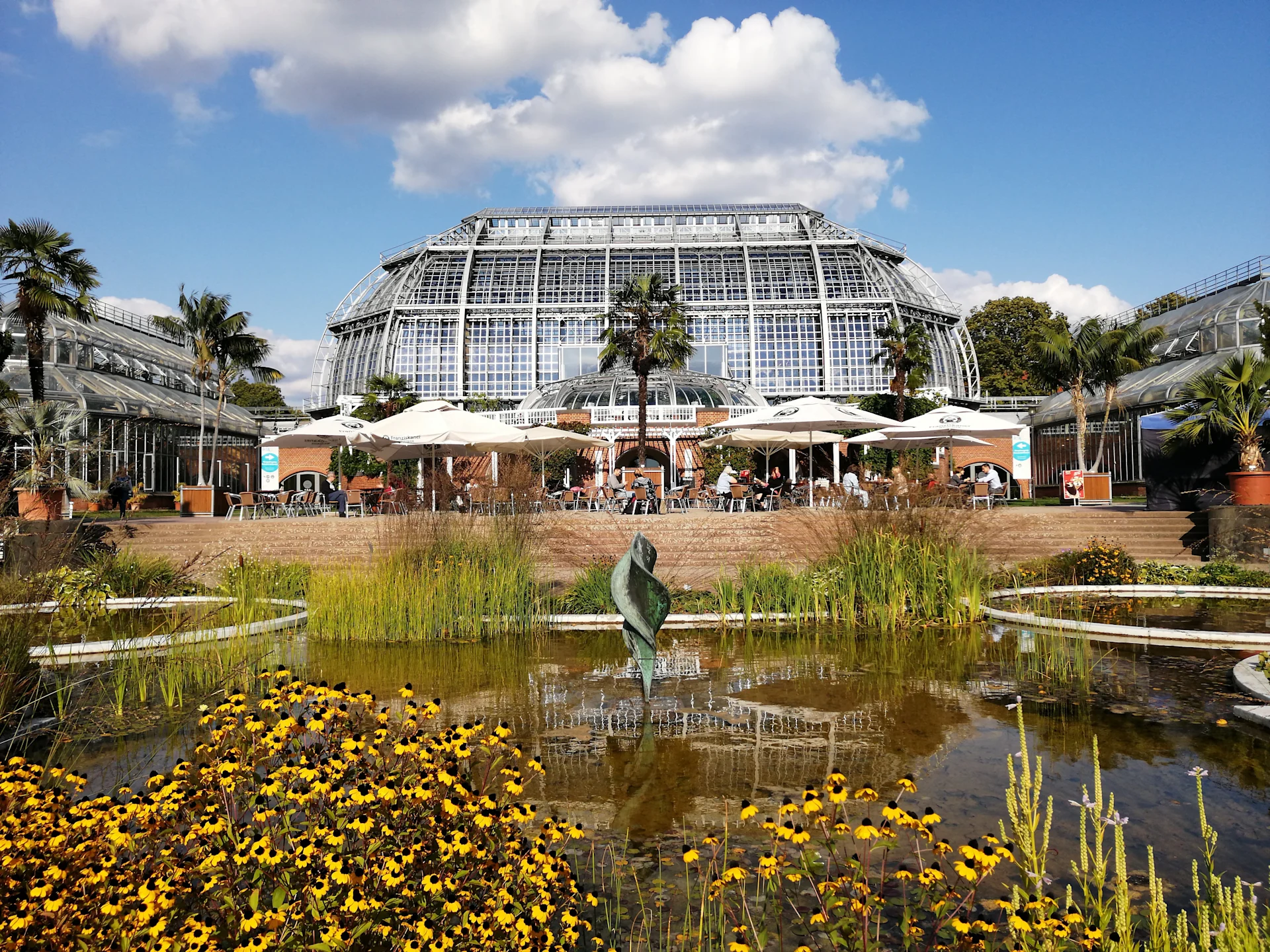 Großes Gewächshaus mit Glasfassade, Teich und Blumen im Vordergrund, Sonnenschirme und Sitzbereich.