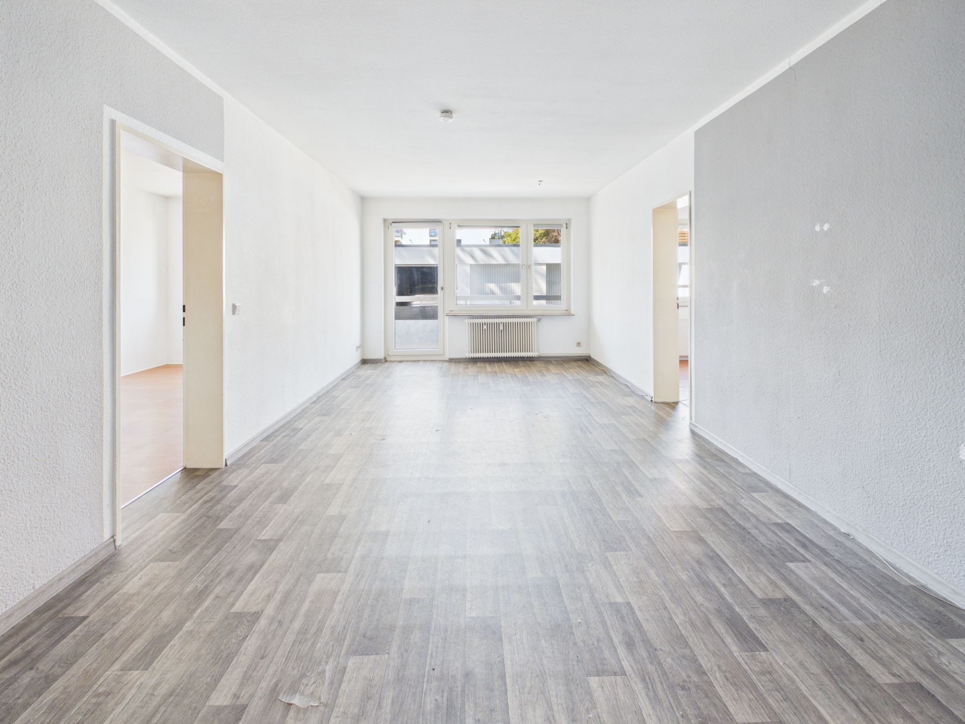 Leeres Zimmer mit großem Fenster, grauem Boden und zwei offenen Türen.