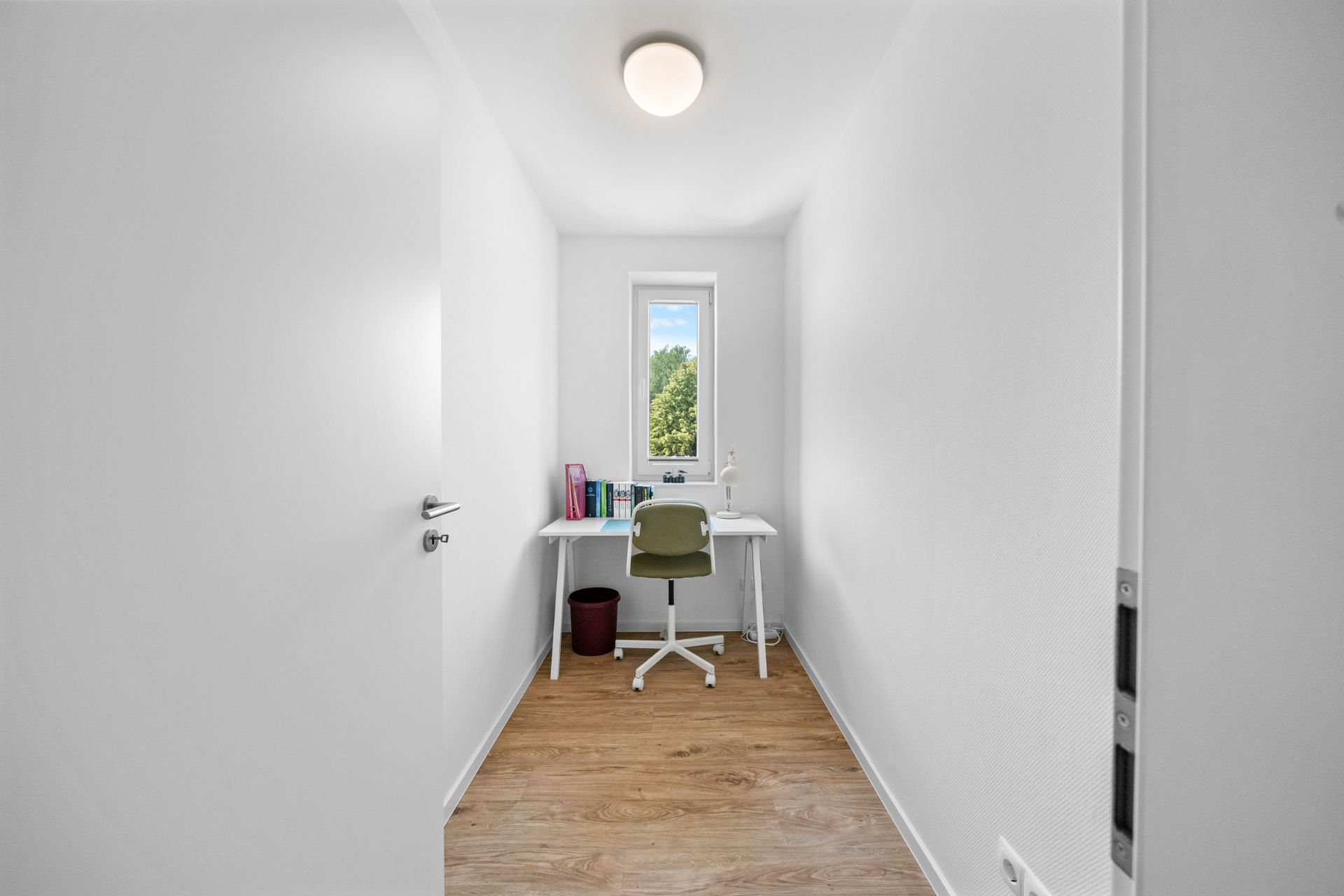 Schmaler Raum mit Schreibtisch, Stuhl, Lampe, Büchern und Fenster am Ende.