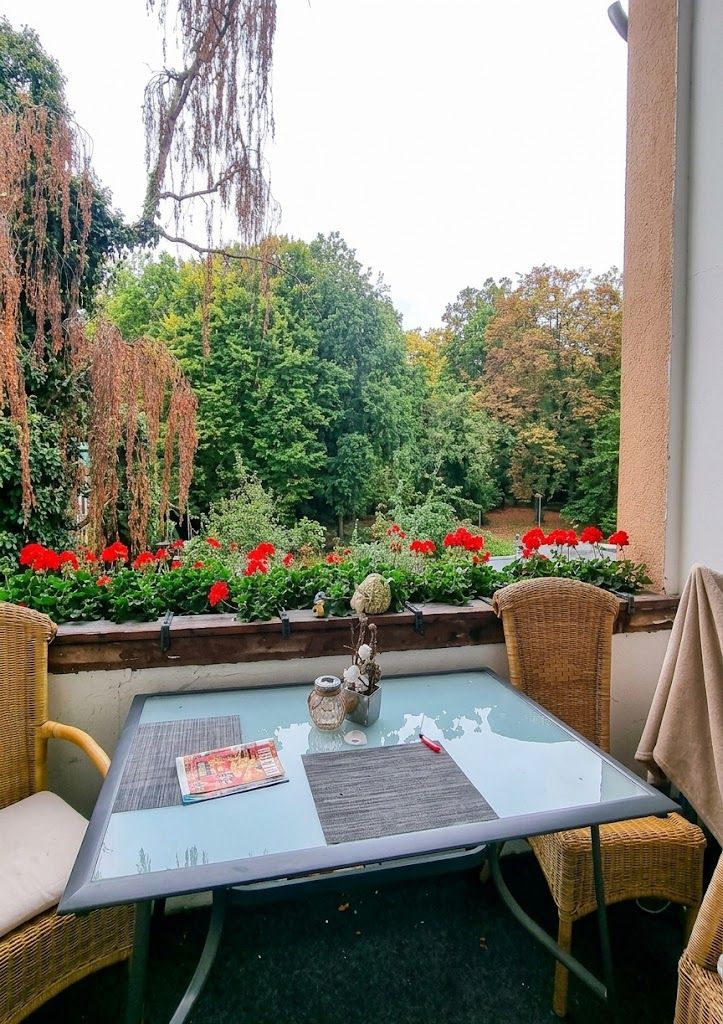 Balkon mit Glastisch, zwei Stühlen und roten Blumen, Blick auf grüne Bäume.