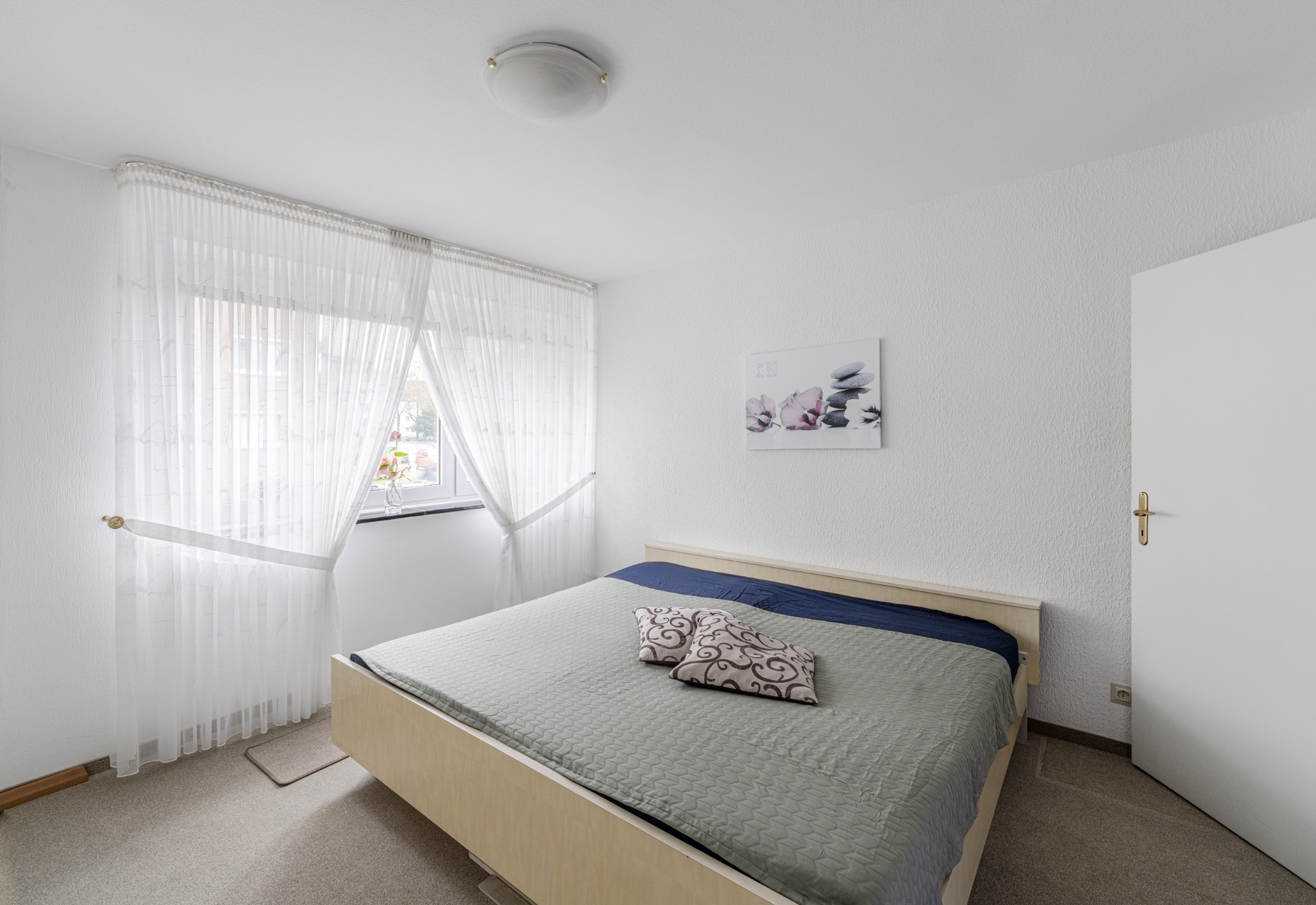 Helles Schlafzimmer mit Doppelbett, zwei Kissen, Fenster mit Vorhängen und Wandbild.