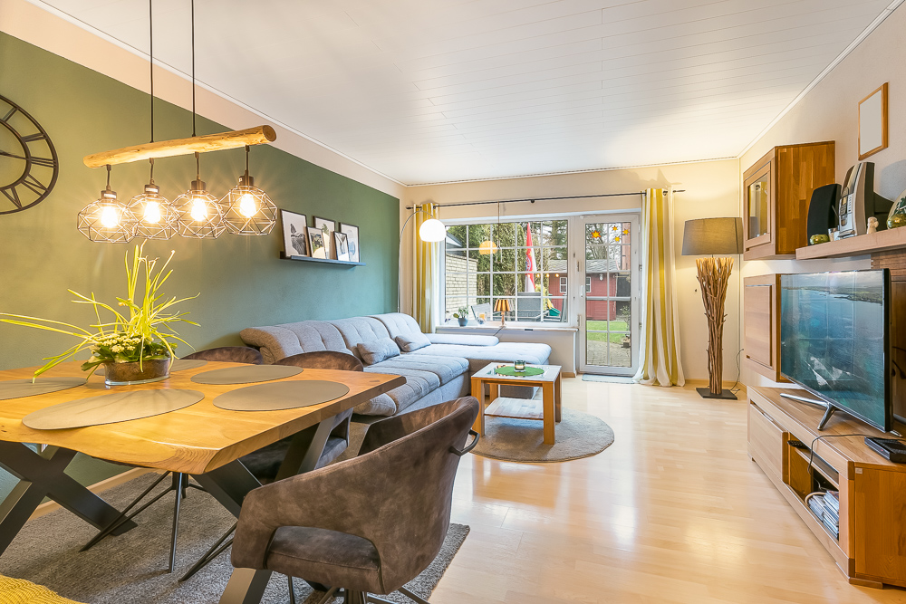 Modernes Wohnzimmer mit grauem Sofa, Holztisch, Fernseher und großen Fenstern zum Garten.