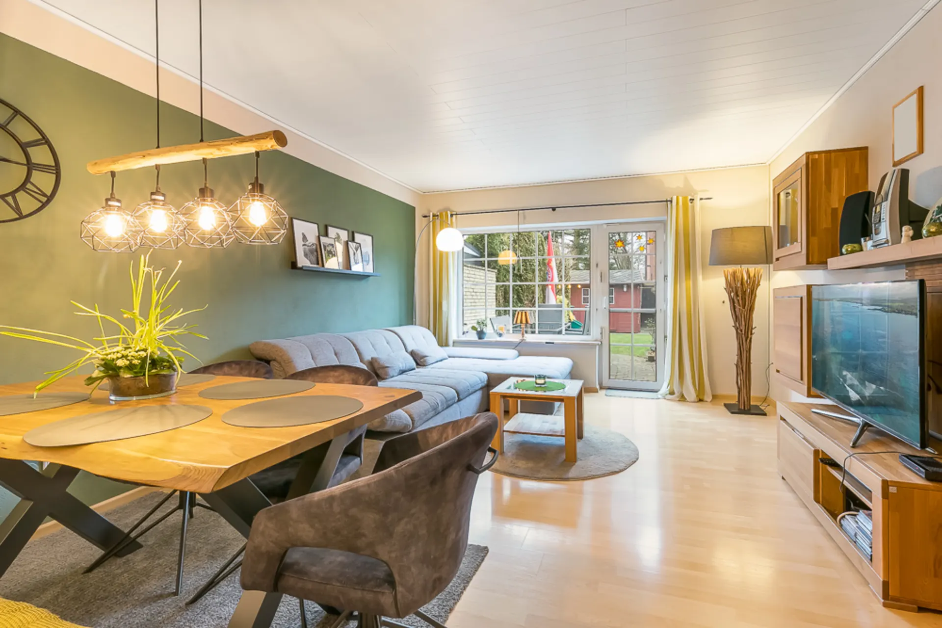 Modernes Wohnzimmer mit grauem Sofa, Holztisch, Fernseher und großen Fenstern zum Garten.