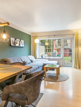 Modernes Wohnzimmer mit grauem Sofa, Holztisch, Fernseher und großen Fenstern zum Garten.