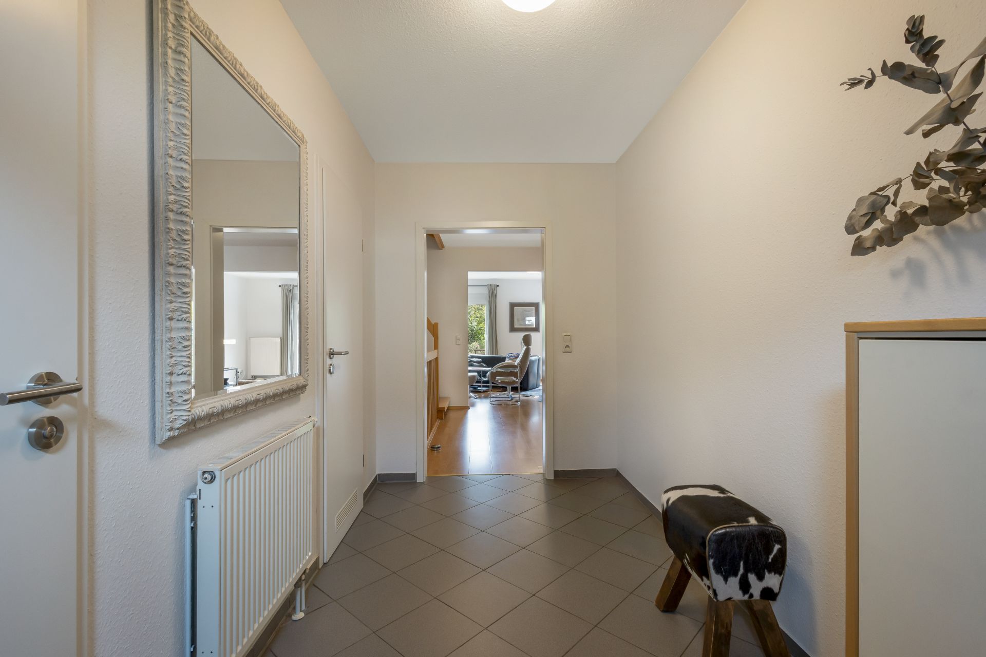 Heller Flur mit Spiegel, kleinem Hocker, Schrank und Blick in ein Wohnzimmer.