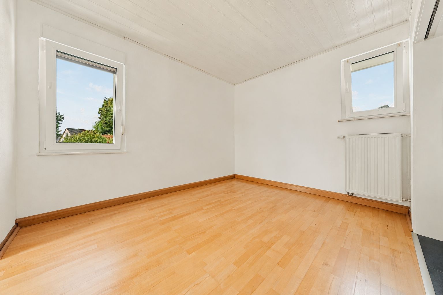 Leeres Zimmer mit Holzfußboden, weißen Wänden und zwei Fenstern.