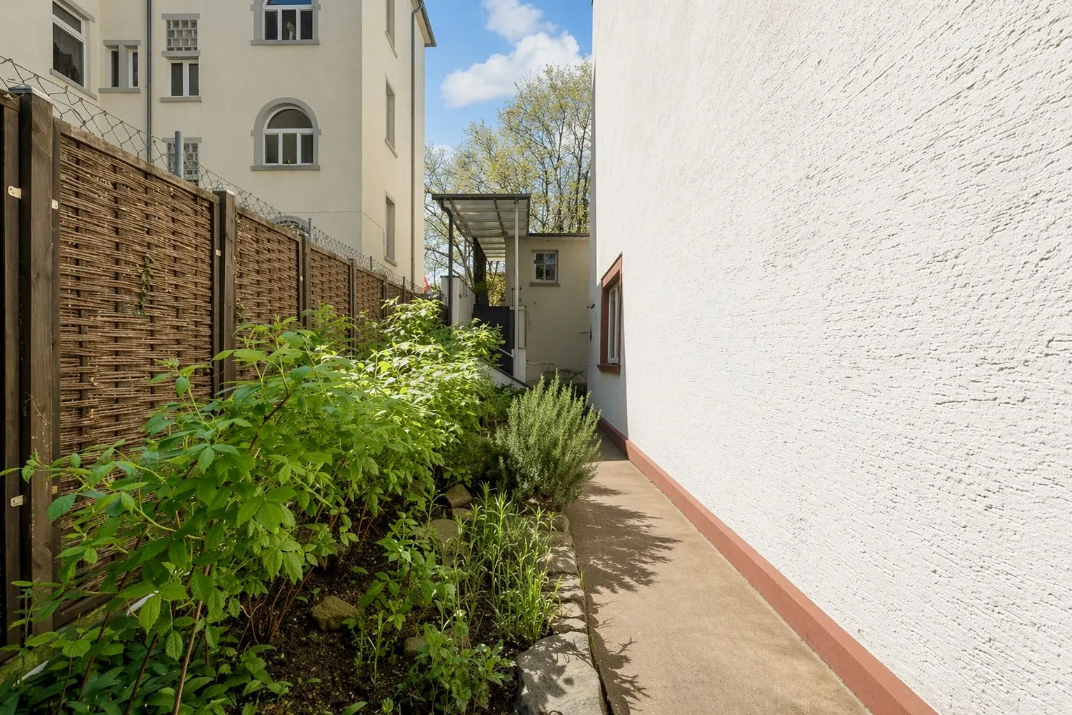 Schmaler Gartenweg mit Pflanzenbeet, Holzzaun und angrenzendem Wohngebäude bei Tageslicht.