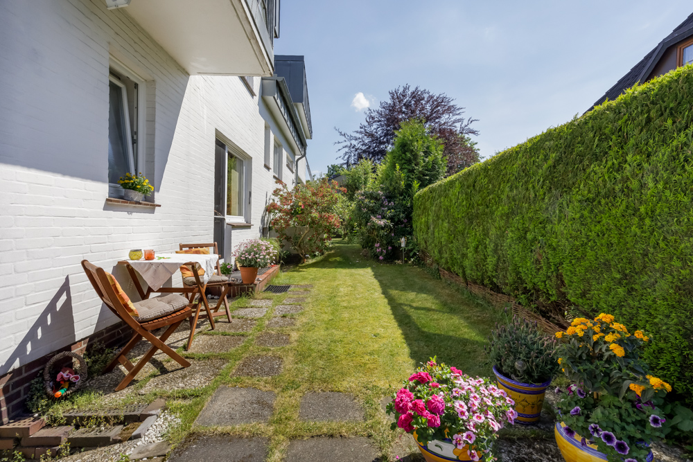 Garten mit Terrasse, Gartenmöbeln und vielen blühenden Blumen neben einem weißen Wohnhaus.