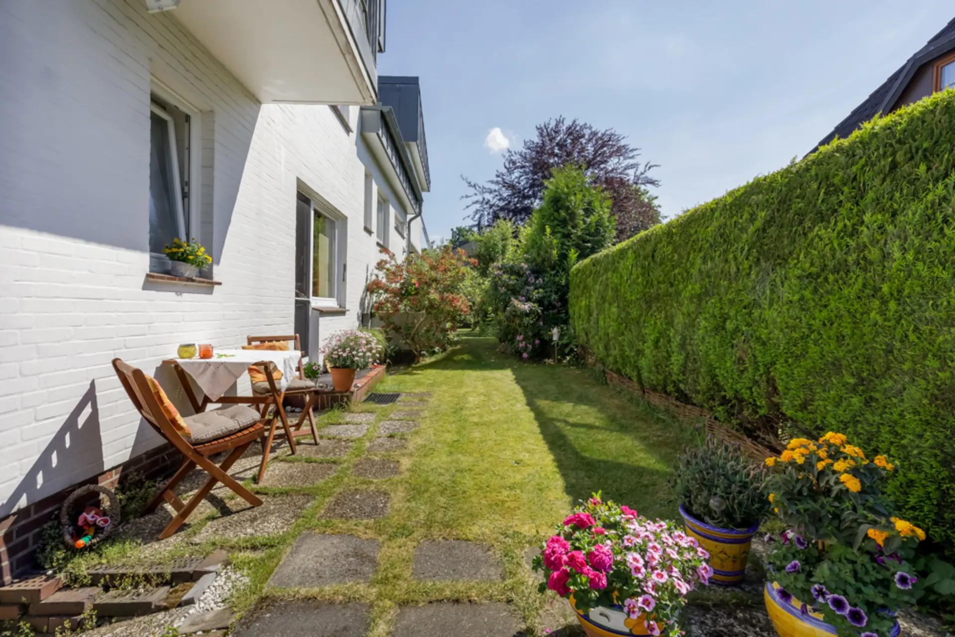 Garten mit Terrasse, Gartenmöbeln und vielen blühenden Blumen neben einem weißen Wohnhaus.
