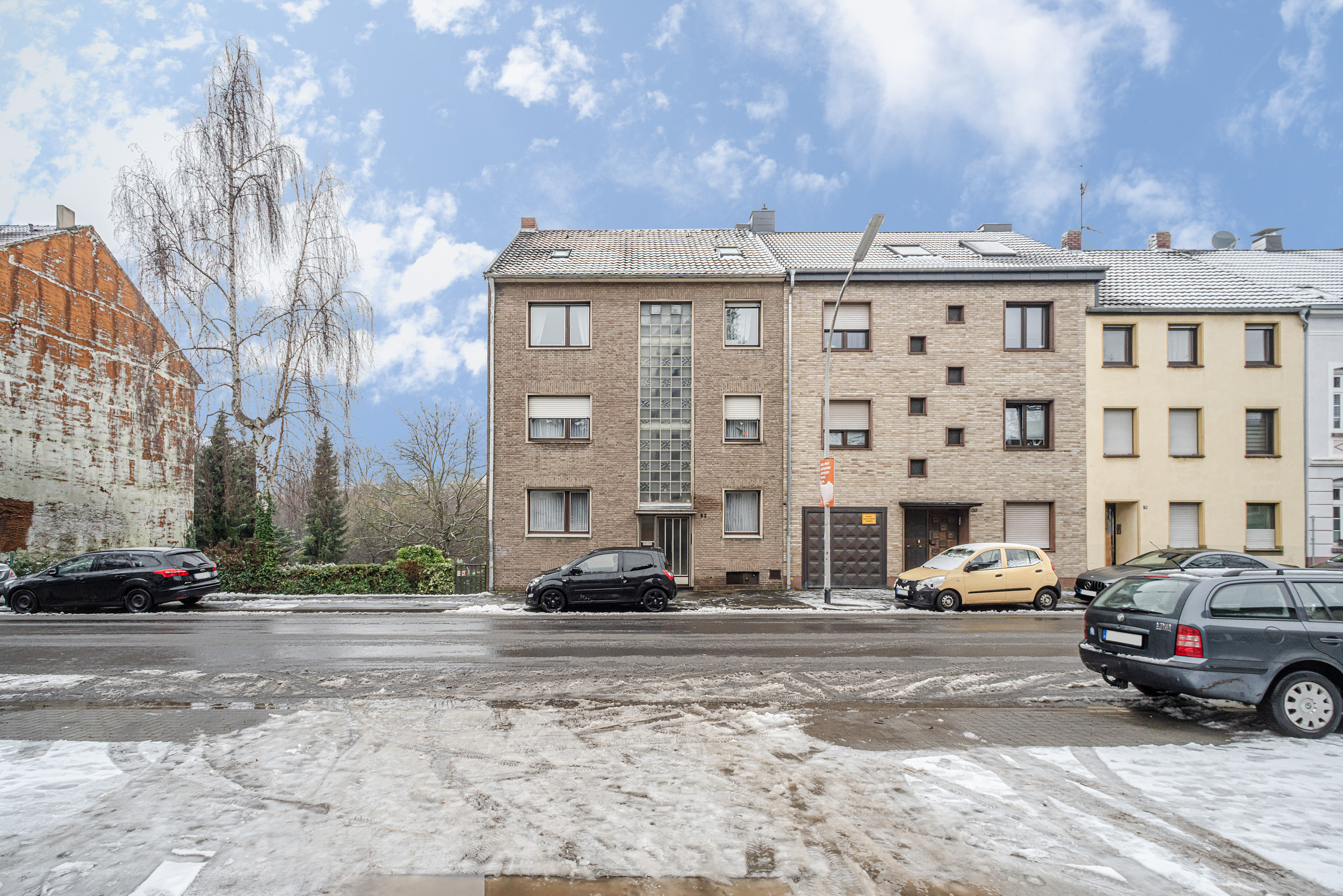 Mehrfamilienhaus an einer verschneiten Straße mit parkenden Autos und Bäumen im Hintergrund.
