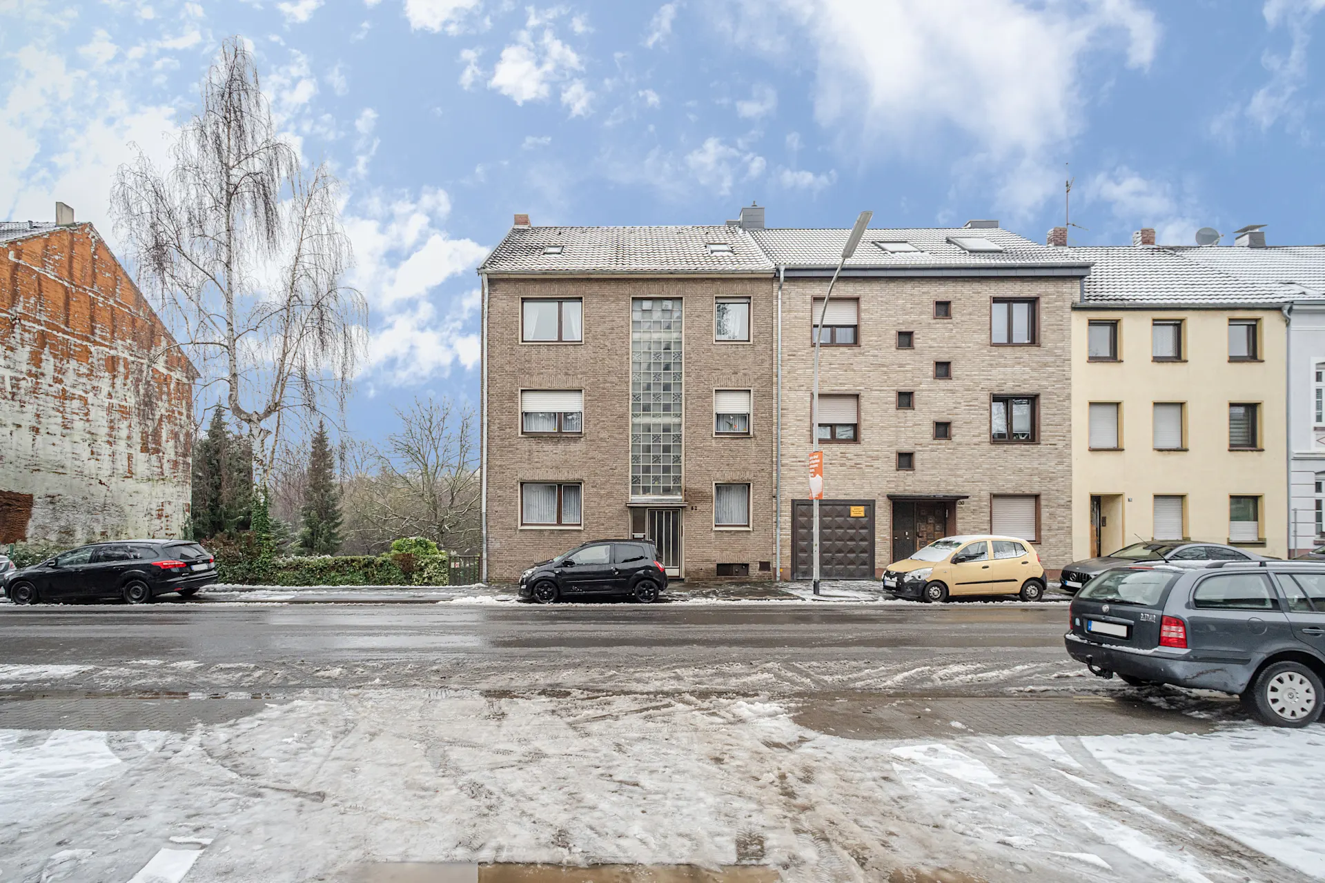 Mehrfamilienhaus an einer verschneiten Straße mit parkenden Autos und Bäumen im Hintergrund.