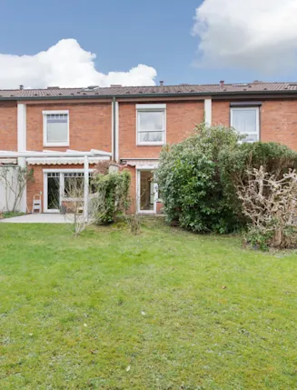 Reihenhaus mit Terrasse und grünem Garten, umgeben von Sträuchern und Bäumen.