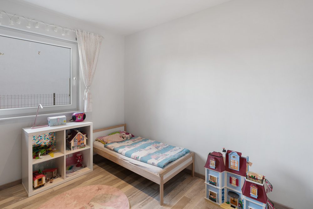 Helles Kinderzimmer mit Einzelbett, Spielzeugregal und Puppenhaus auf Holzboden.
