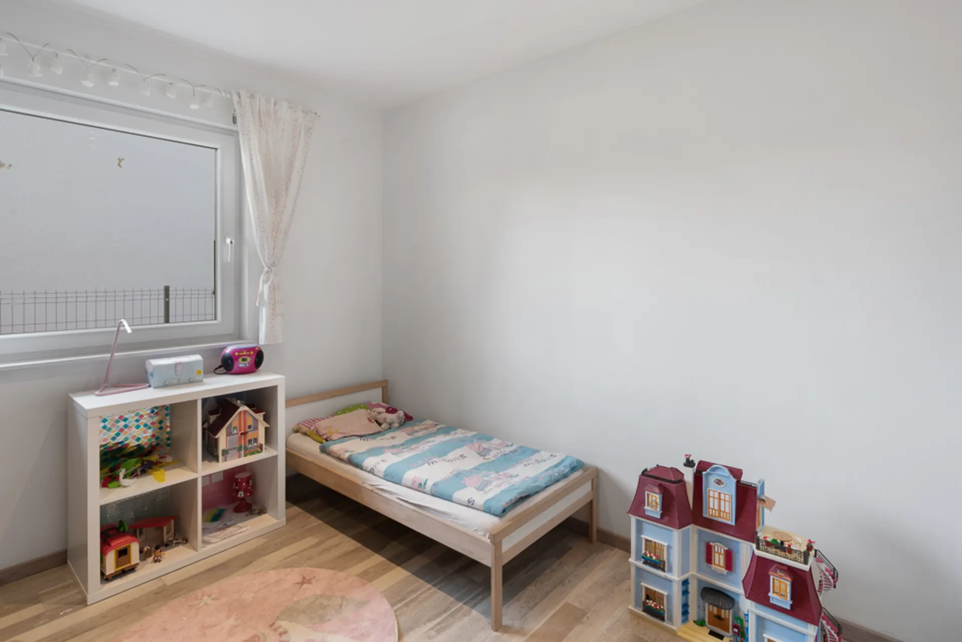 Helles Kinderzimmer mit Einzelbett, Spielzeugregal und Puppenhaus auf Holzboden.