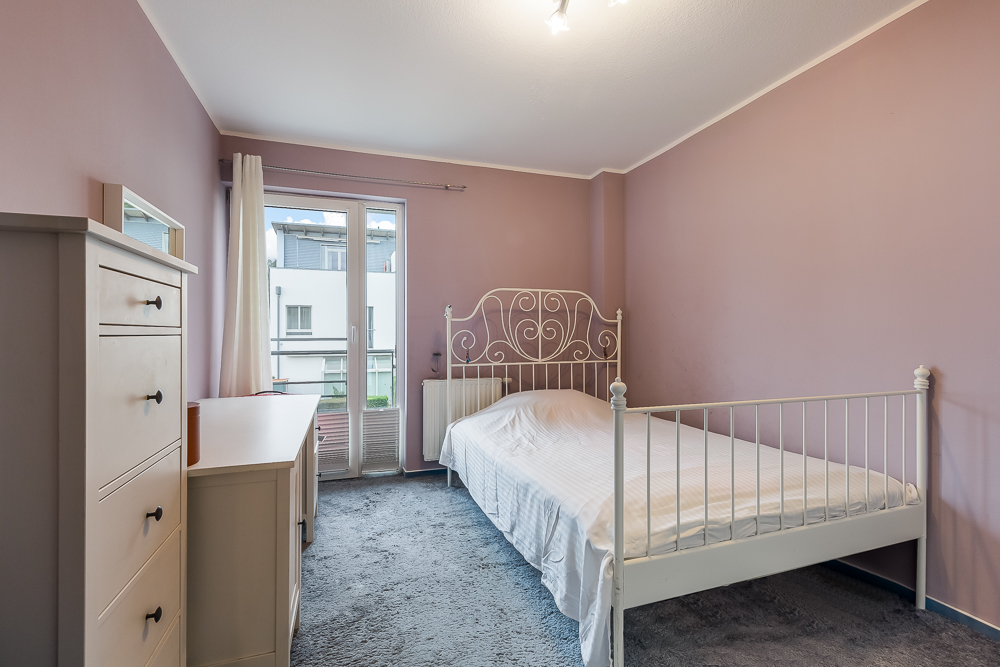 Schlafzimmer mit weißem Bett, Kommode, Schreibtisch und rosa Wänden, Fenster zur Terrasse.