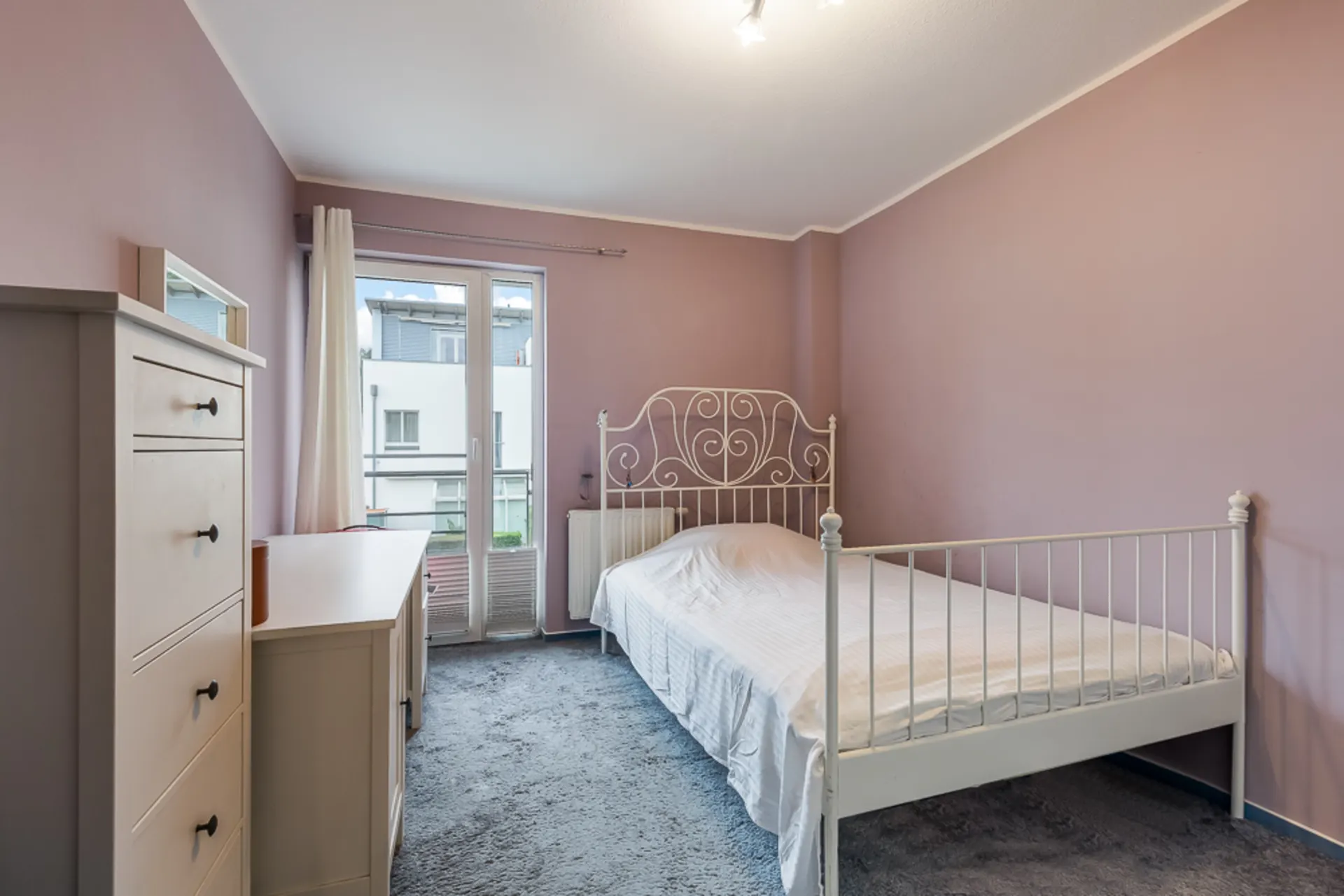 Schlafzimmer mit weißem Bett, Kommode, Schreibtisch und rosa Wänden, Fenster zur Terrasse.