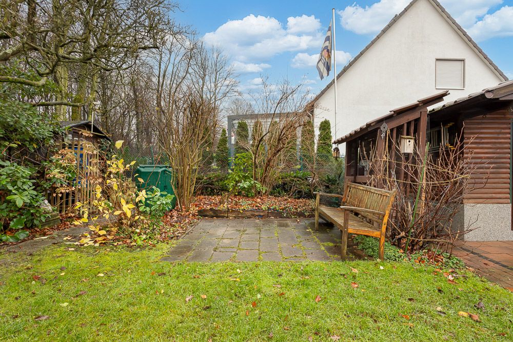 Garten mit Steinplatten, Holzbank, Schuppen, Rasenfläche und Haus im Hintergrund.