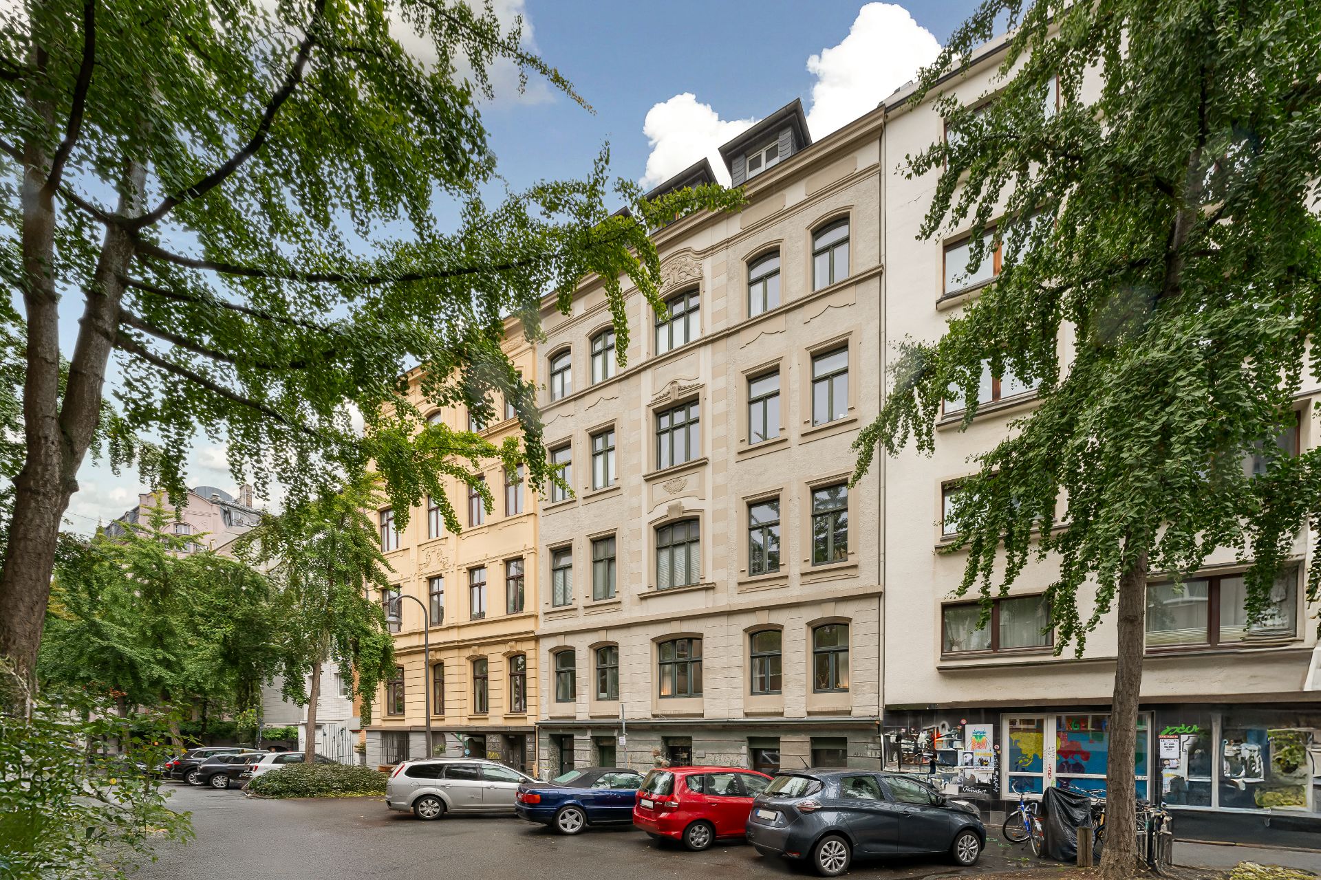 Mehrstöckiges Wohnhaus mit Bäumen und geparkten Autos in einer ruhigen Straße.
