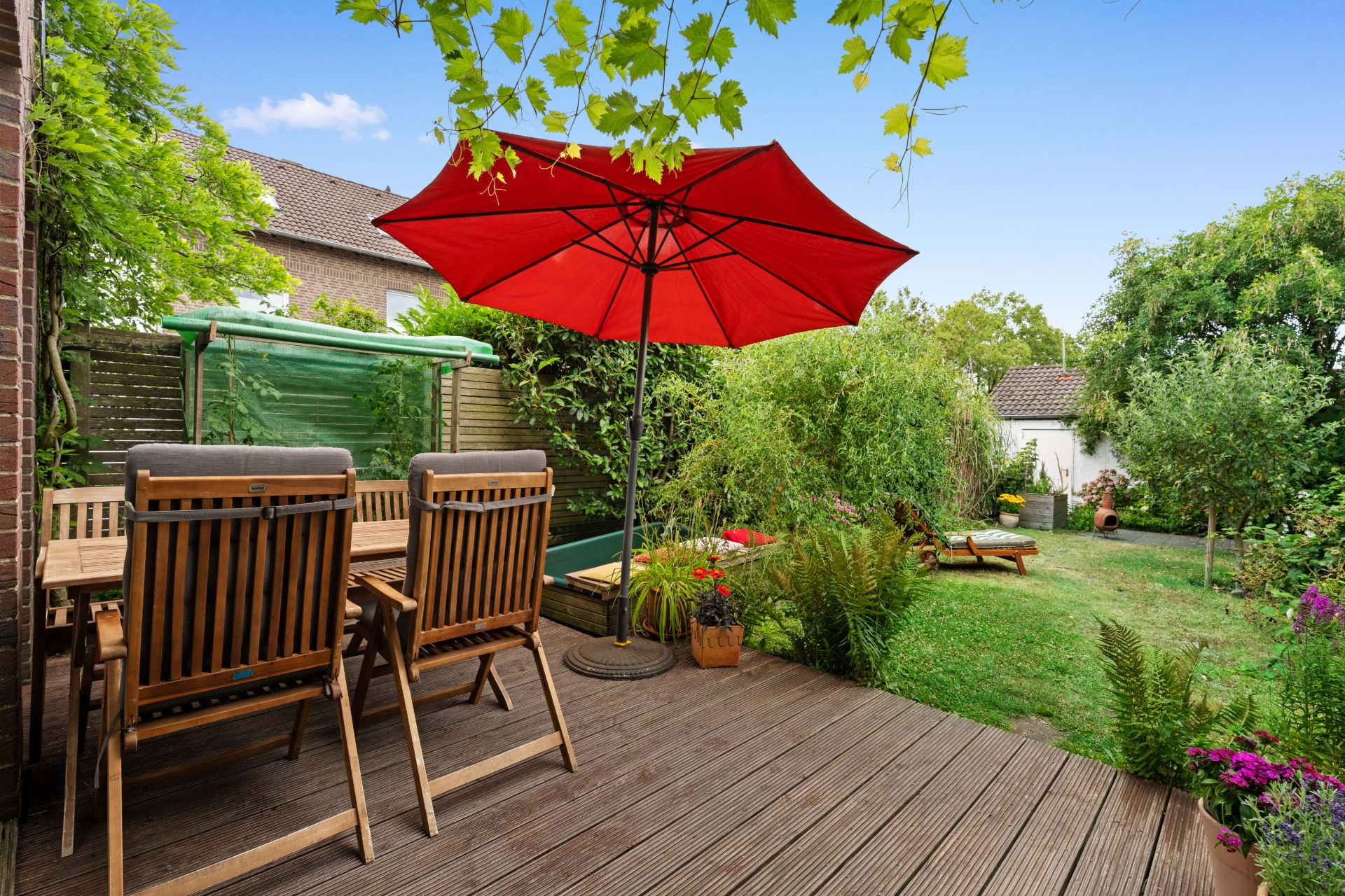 Holzterrasse mit Tisch, Stühlen und rotem Sonnenschirm, Blick auf Garten mit Liegestuhl und Pflanzen.