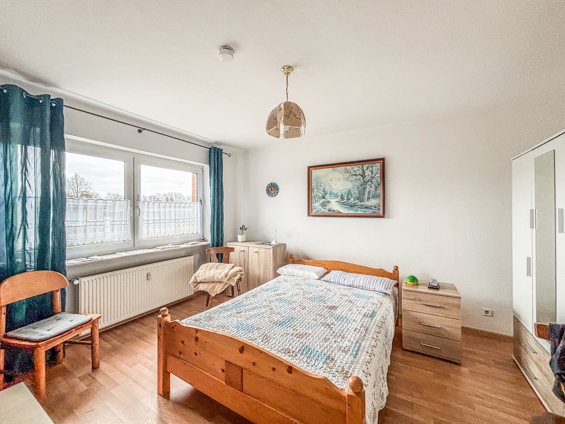 Helles Schlafzimmer mit Holzbett, Schrank, Stühlen und großem Fenster mit Vorhängen.