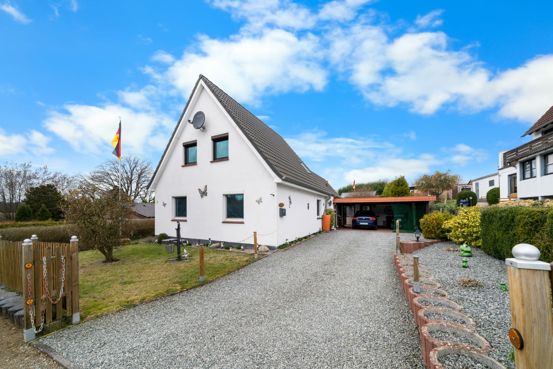 Weißes Einfamilienhaus mit spitzem Dach, Auffahrt und gepflegtem Garten bei blauem Himmel.