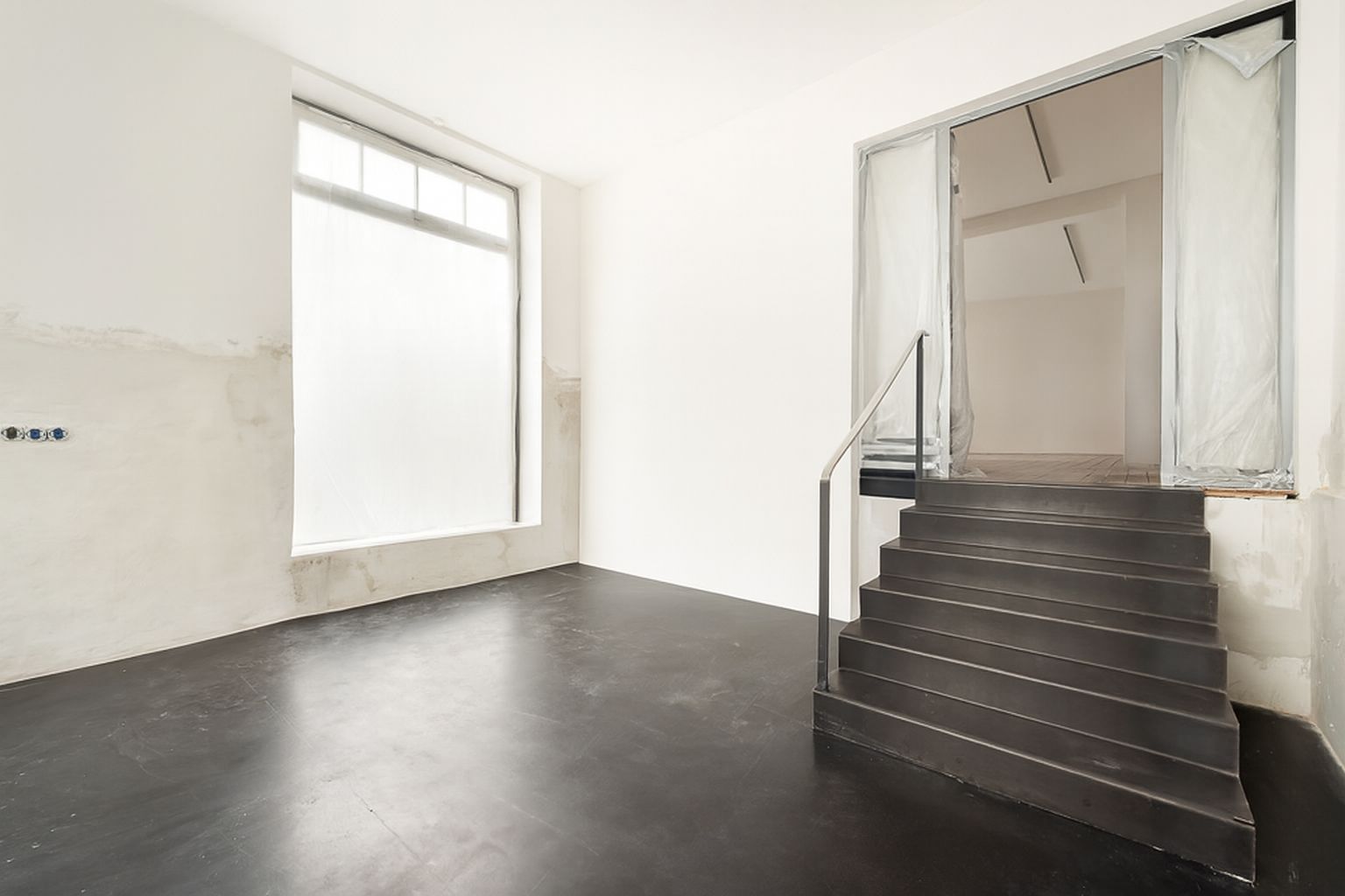 Heller Raum mit großem Fenster und schwarzer Treppe, die zu einer erhöhten Ebene führt.
