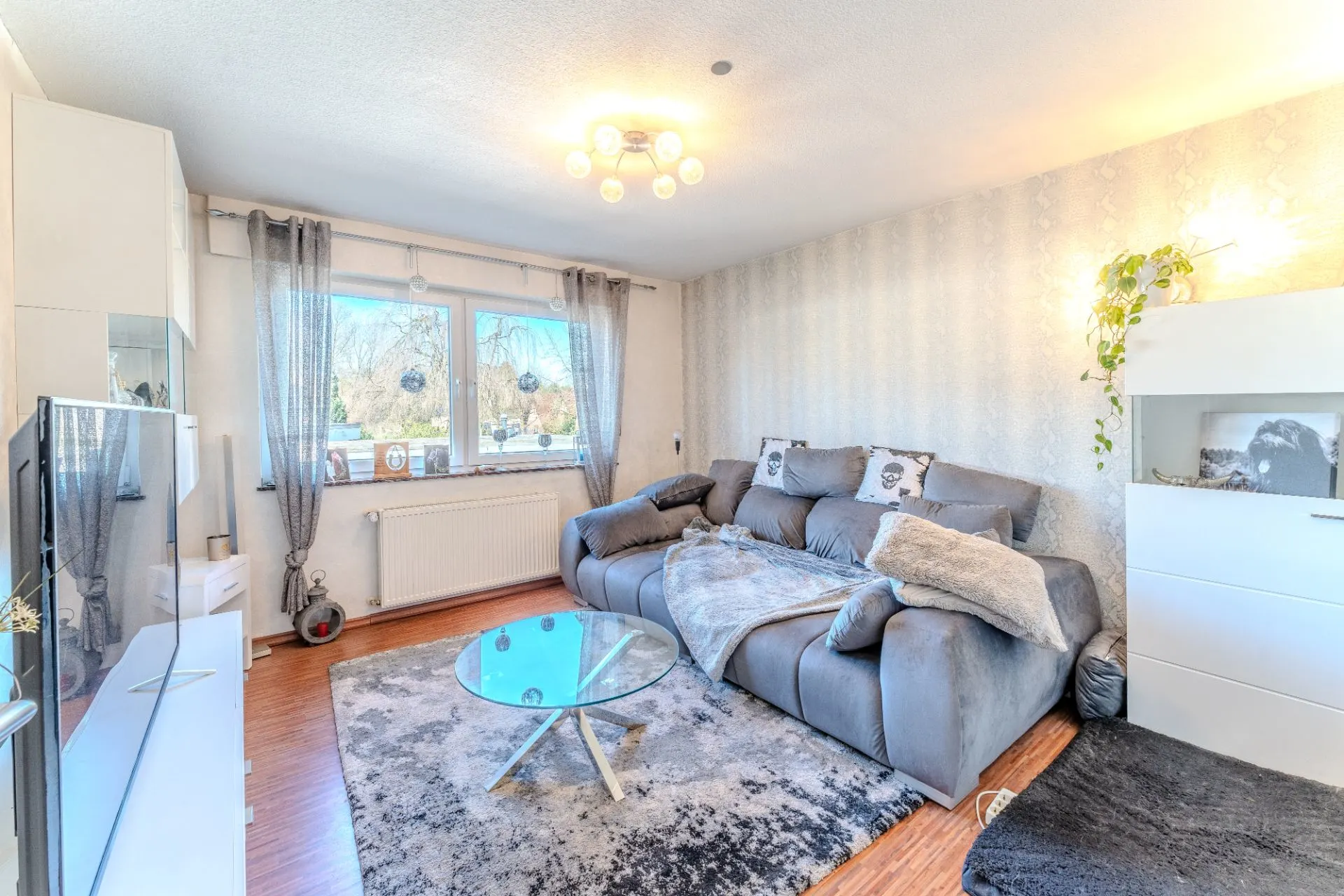 Helles Wohnzimmer mit grauem Sofa, Glastisch, Teppich und großem Fenster.