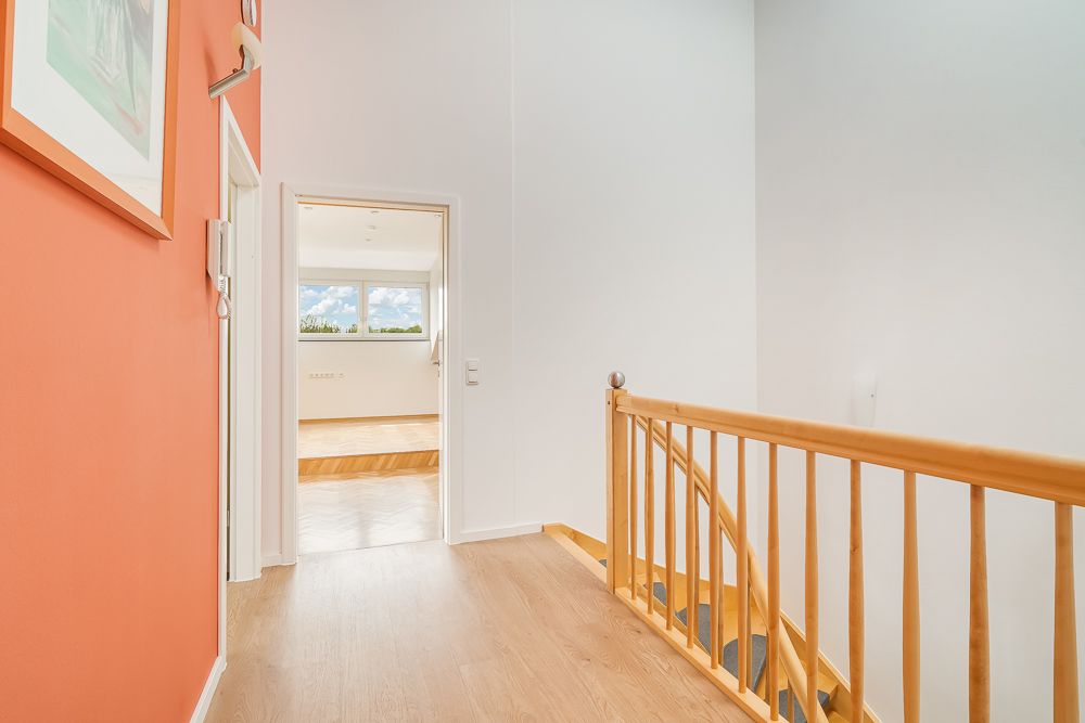 Heller Flur mit Holztreppe, orangener Wand und Blick in ein Zimmer mit großen Fenstern.