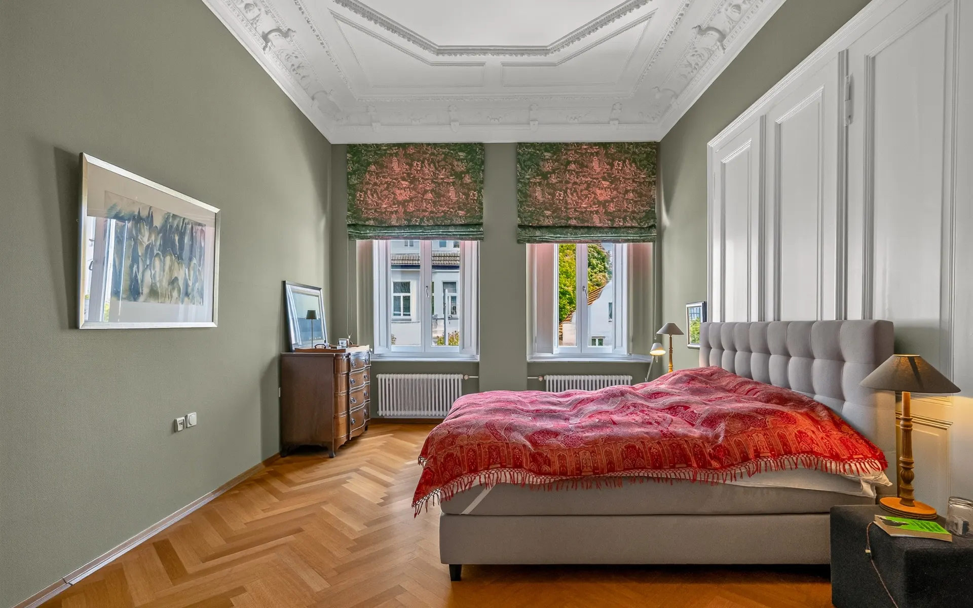 Schlafzimmer mit großem Bett, Parkettboden und zwei Fenstern mit grünen Vorhängen.