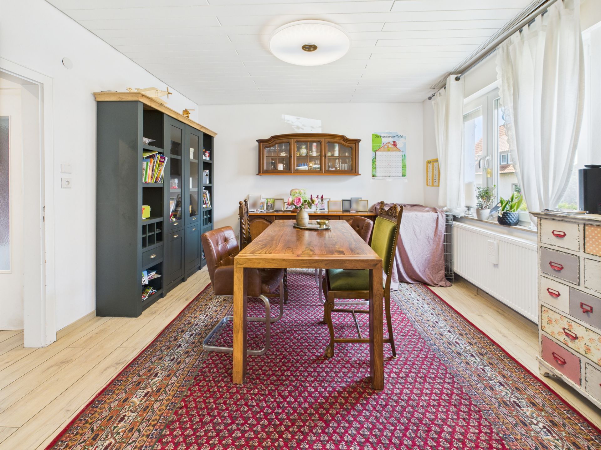 Esszimmer mit Holztisch, gemischten Stühlen, Teppich und Regal an der Wand.