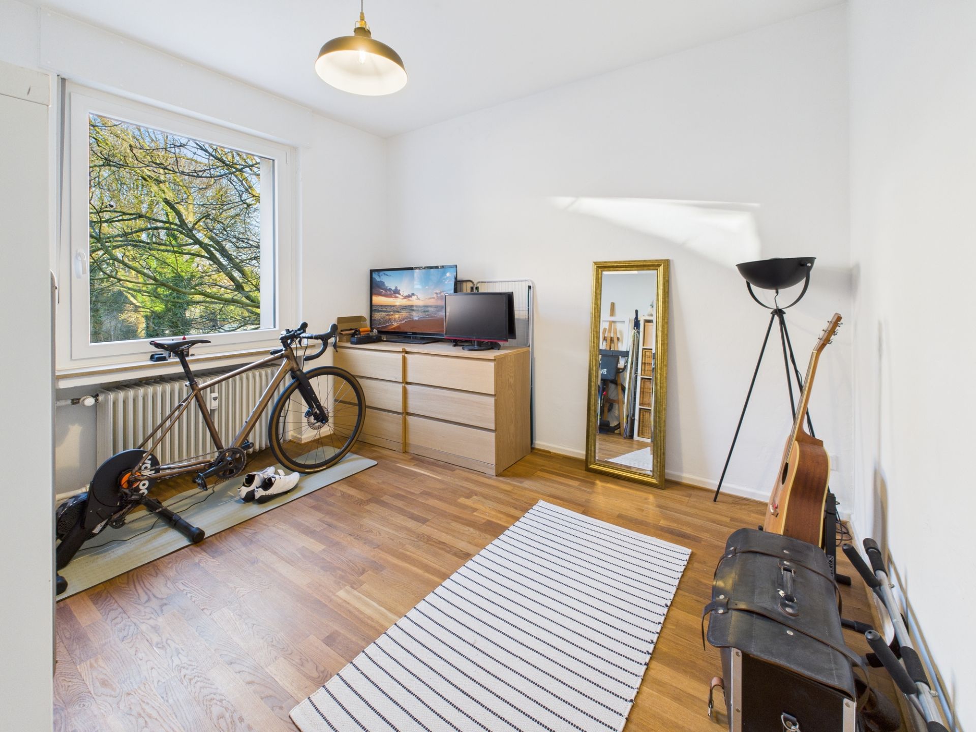 Helles Zimmer mit Fahrrad, Kommode, Fernseher, Spiegel, Stehlampe, Gitarre und Teppich.