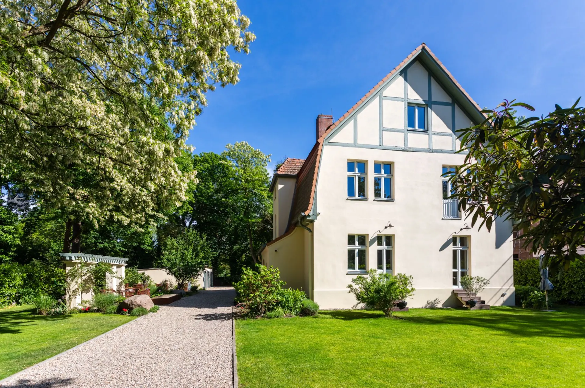 Helles Wohnhaus mit gepflegtem Garten, Kiesweg und Bäumen an einem sonnigen Tag.