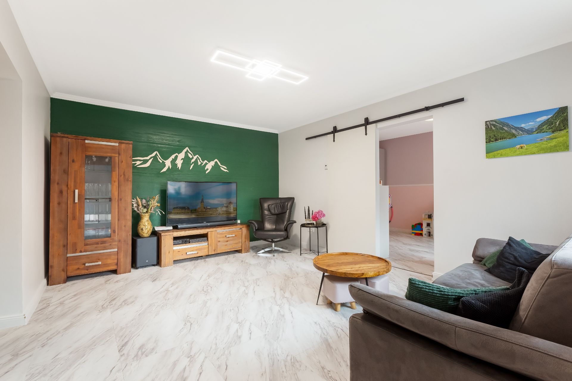 Modernes Wohnzimmer mit Sofa, Holzmöbeln und grün gestrichener Wand mit Bergmotiv.