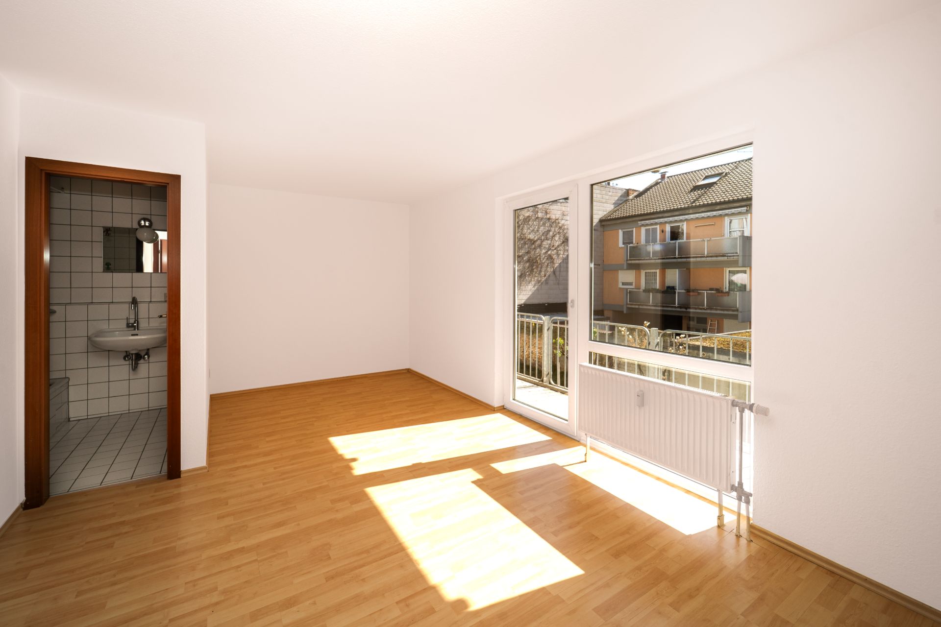 Helles, leeres Zimmer mit Parkettboden, großem Fenster und Zugang zu einem kleinen Bad.