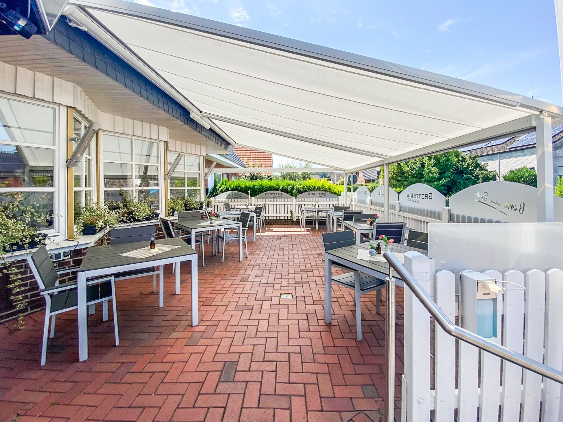 Große Terrasse mit Tischen, Stühlen, Markise und weißen Holzzäunen neben einem Haus.