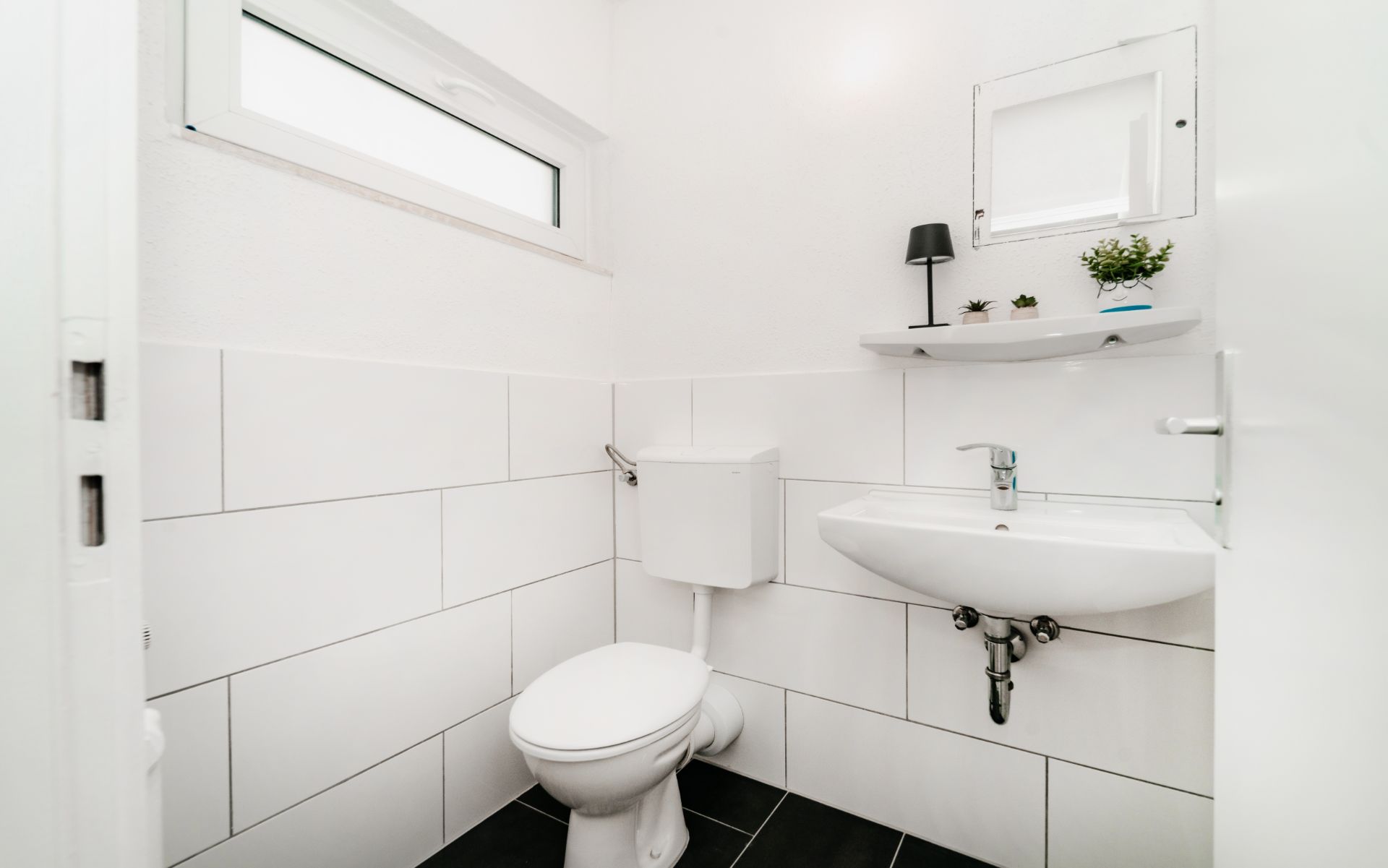 Helles Badezimmer mit WC, Waschbecken, kleinem Fenster und Regal mit Dekoration.