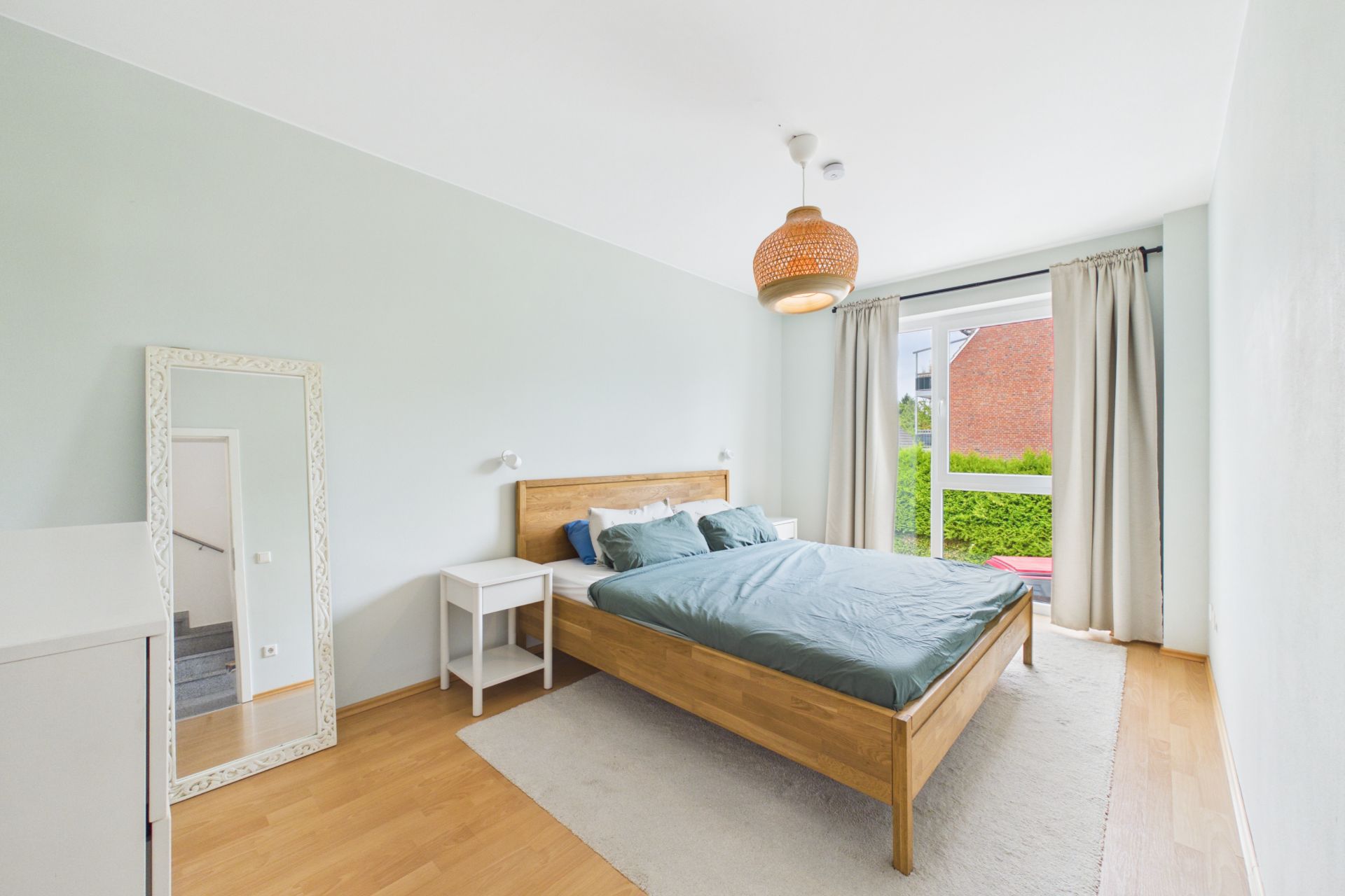 Helles Schlafzimmer mit Holzbett, großem Fenster, Spiegel und Nachttisch.