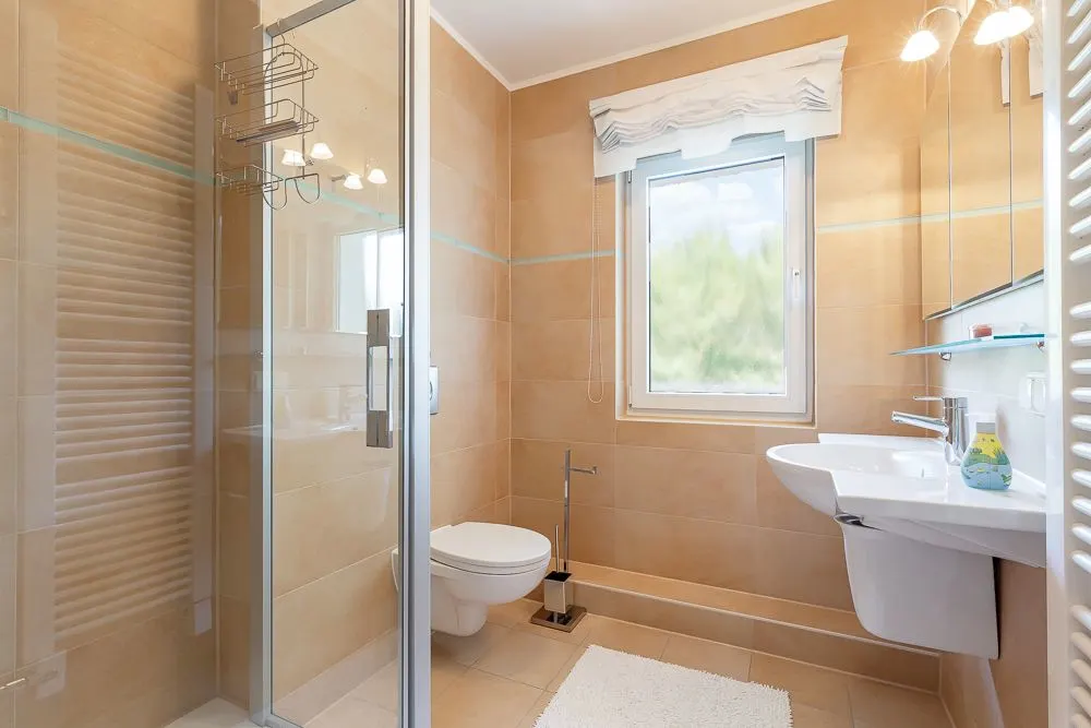 Modernes Badezimmer mit Dusche, WC, Fenster, Waschbecken und hellen Fliesen.