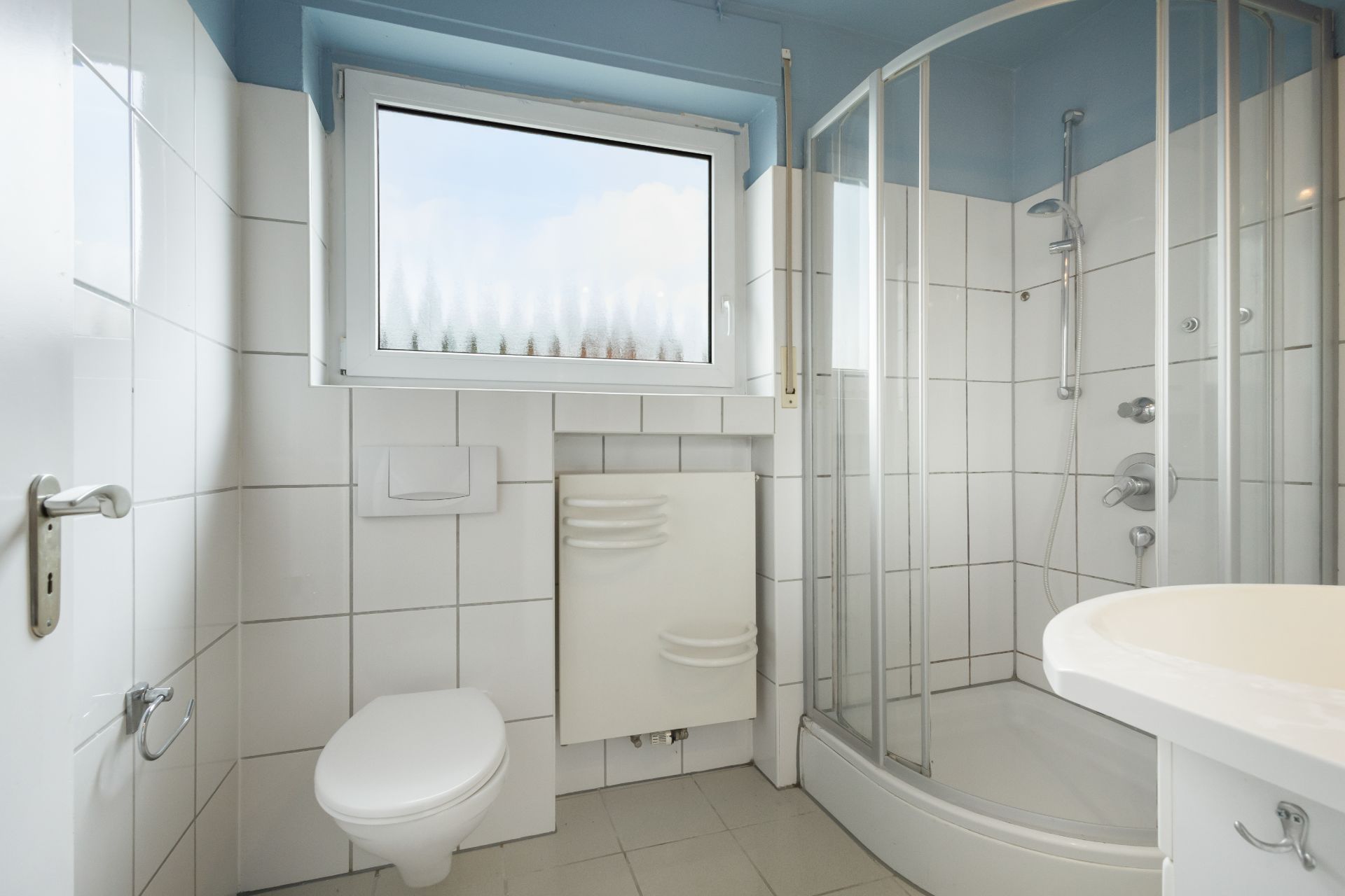 Helles Badezimmer mit Dusche, WC, Fenster und Waschbecken, weiße Fliesen an Wand und Boden.