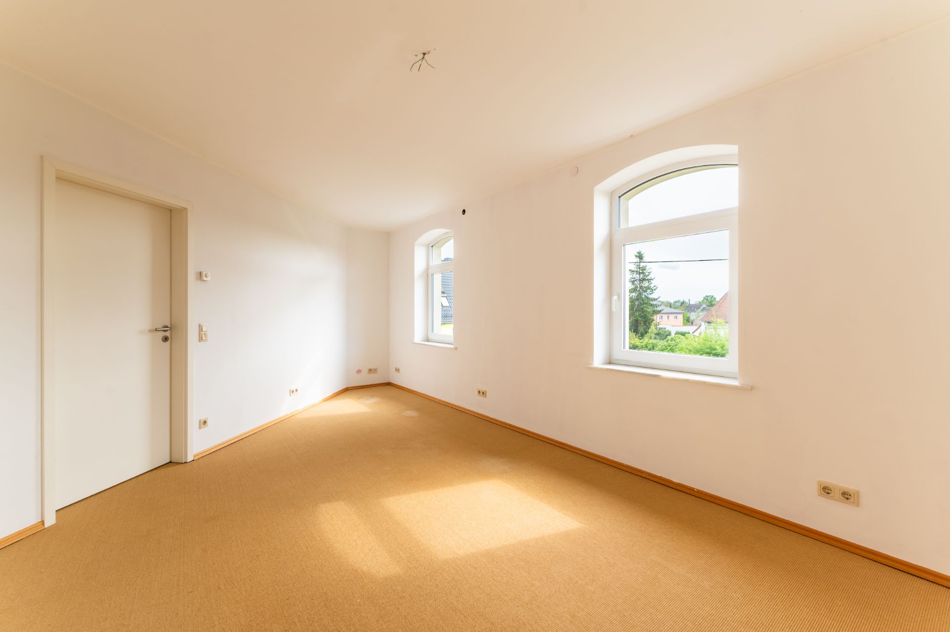 Heller, leerer Raum mit zwei Fenstern, weißer Tür und beigem Teppichboden.