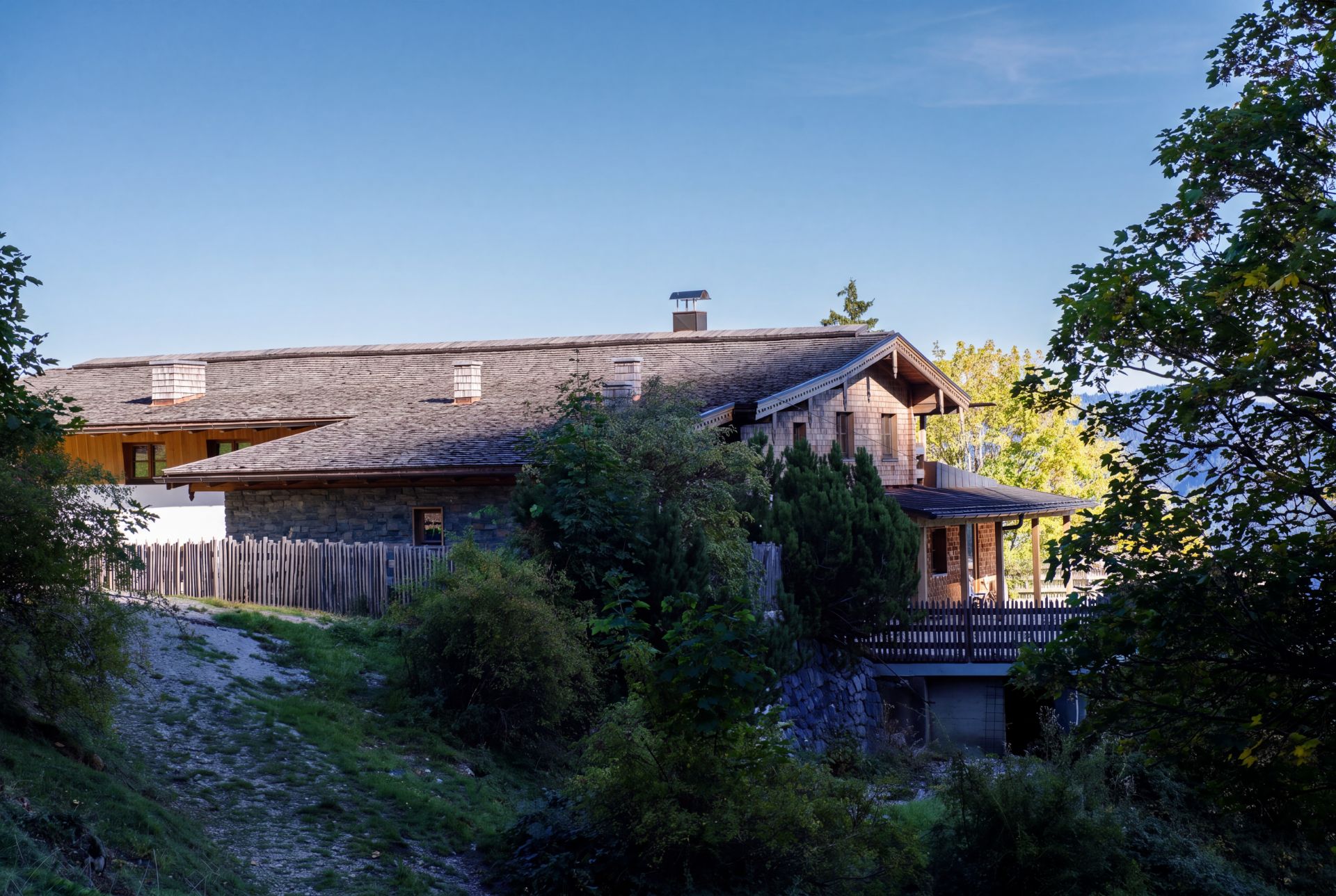 Großes Holzhaus mit Veranda, umgeben von Bäumen und einem Holzzaun, bei blauem Himmel.