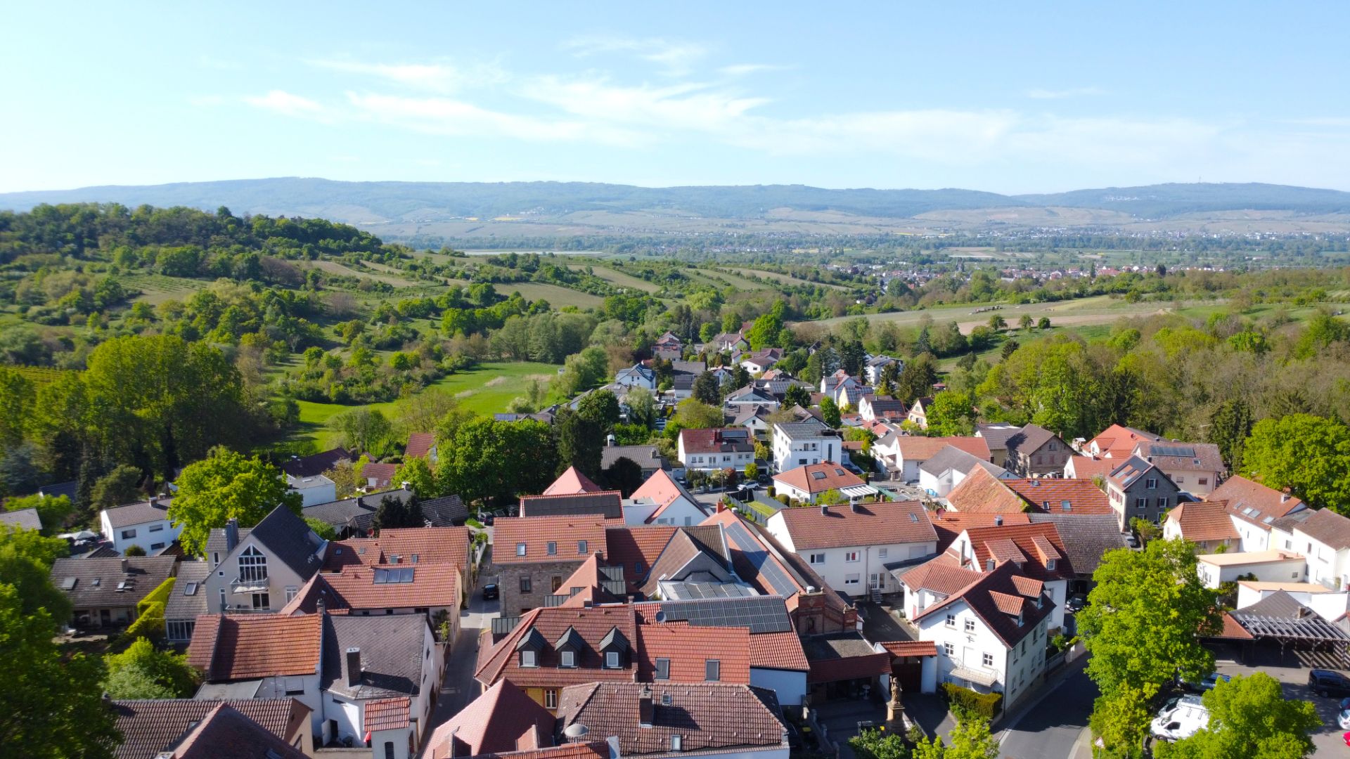 Luftaufnahme eines Wohngebiets mit roten Dächern und viel Grün in hügeliger Landschaft.