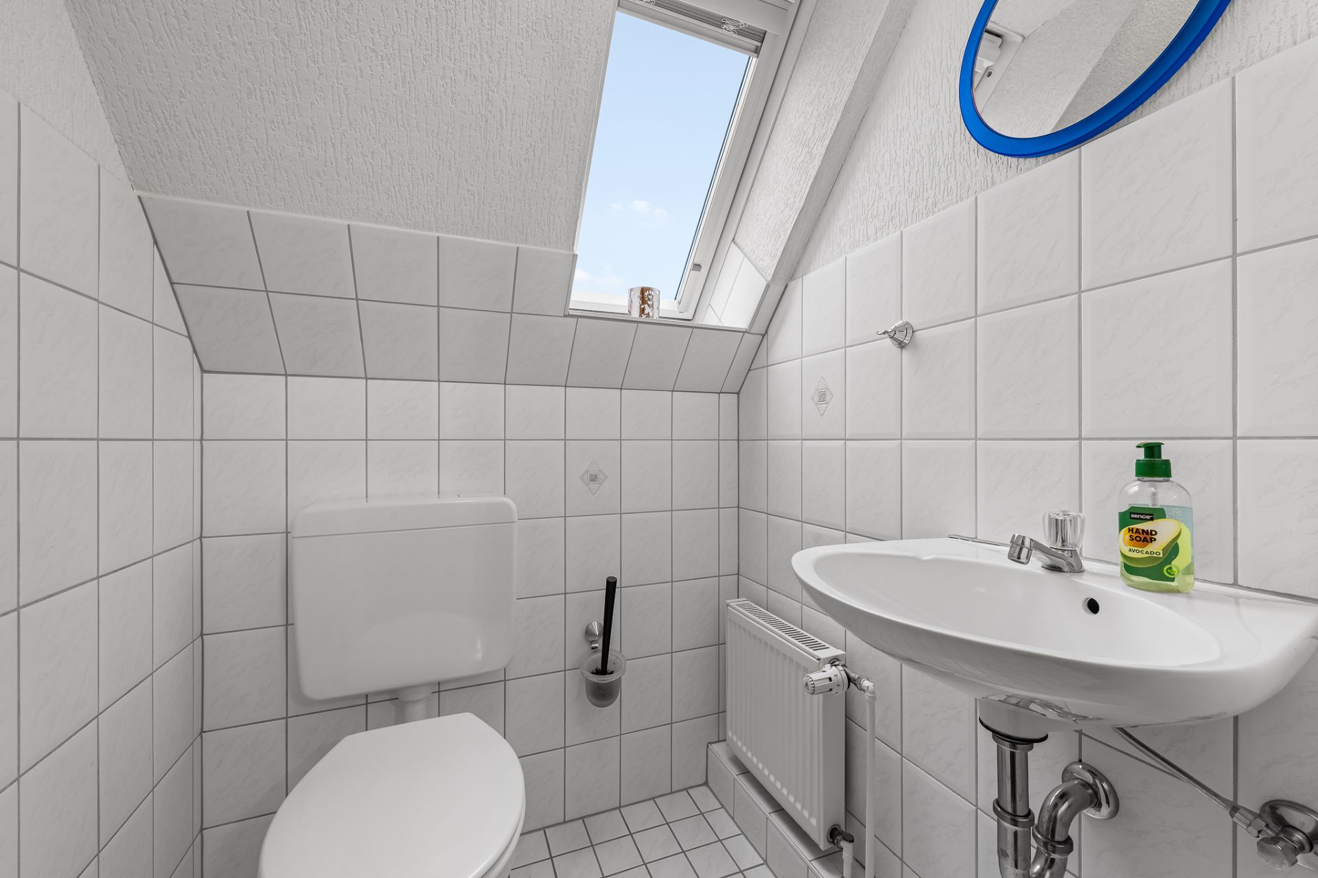 Kleines Badezimmer mit WC, Waschbecken, Spiegel und Dachfenster, weiß gefliest.