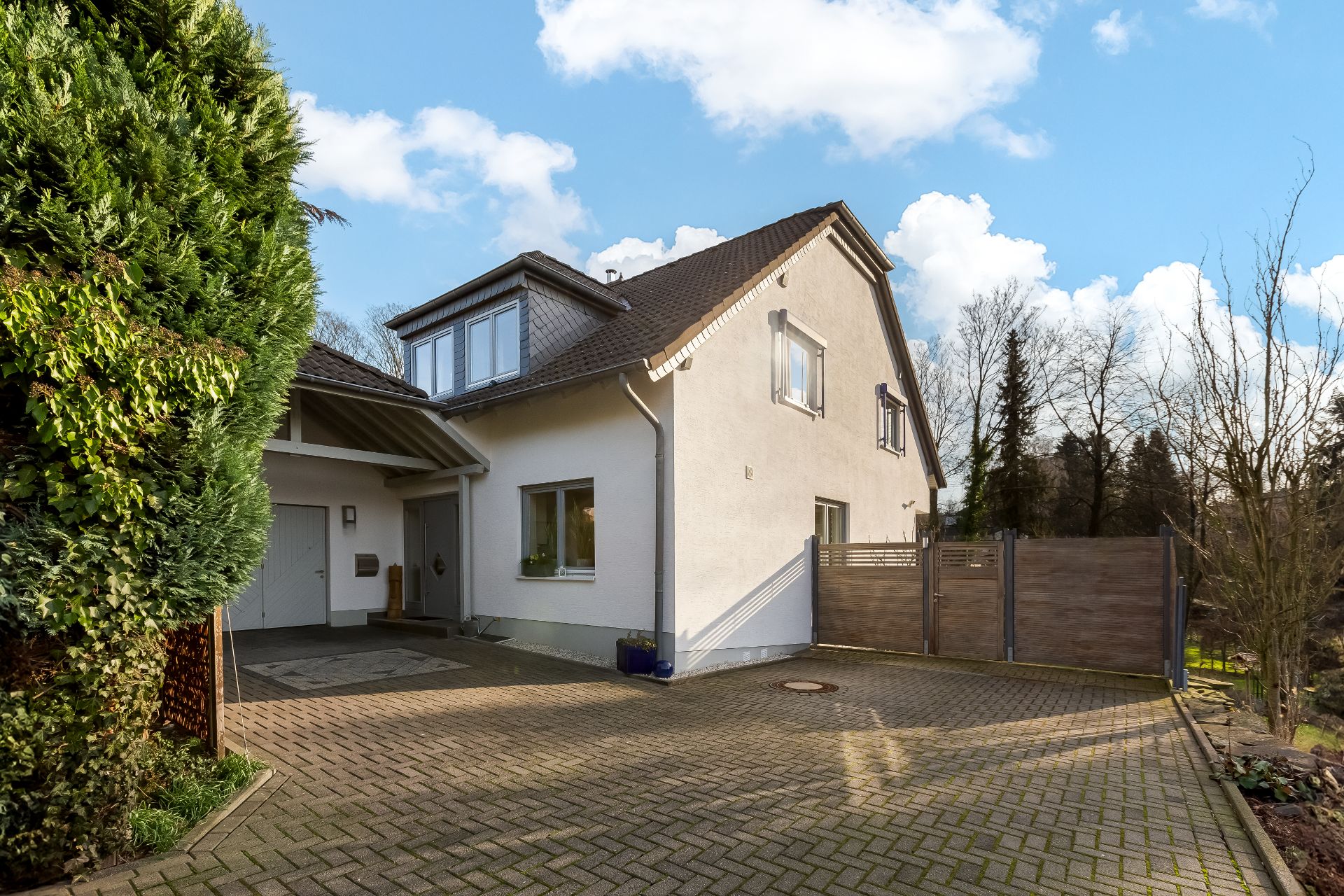 Modernes Einfamilienhaus mit gepflasterter Einfahrt und Garten, links große grüne Hecke.