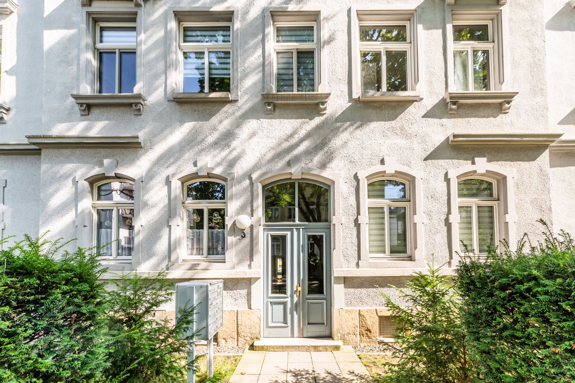 Fassade eines Wohngebäudes mit mehreren Fenstern und einer Haustür, umgeben von Büschen.